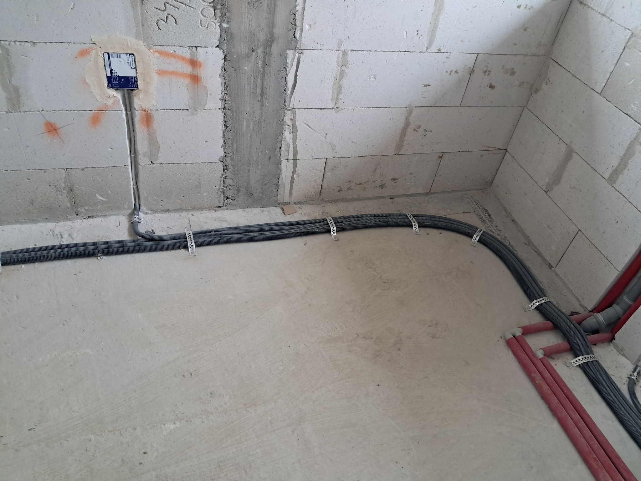 Szare rury w karbowanej osłonie przymocowane do ściany z białych bloczków, z widoczną puszką instalacyjną i fragmentem czerwonych rur w rogu pomieszczenia.