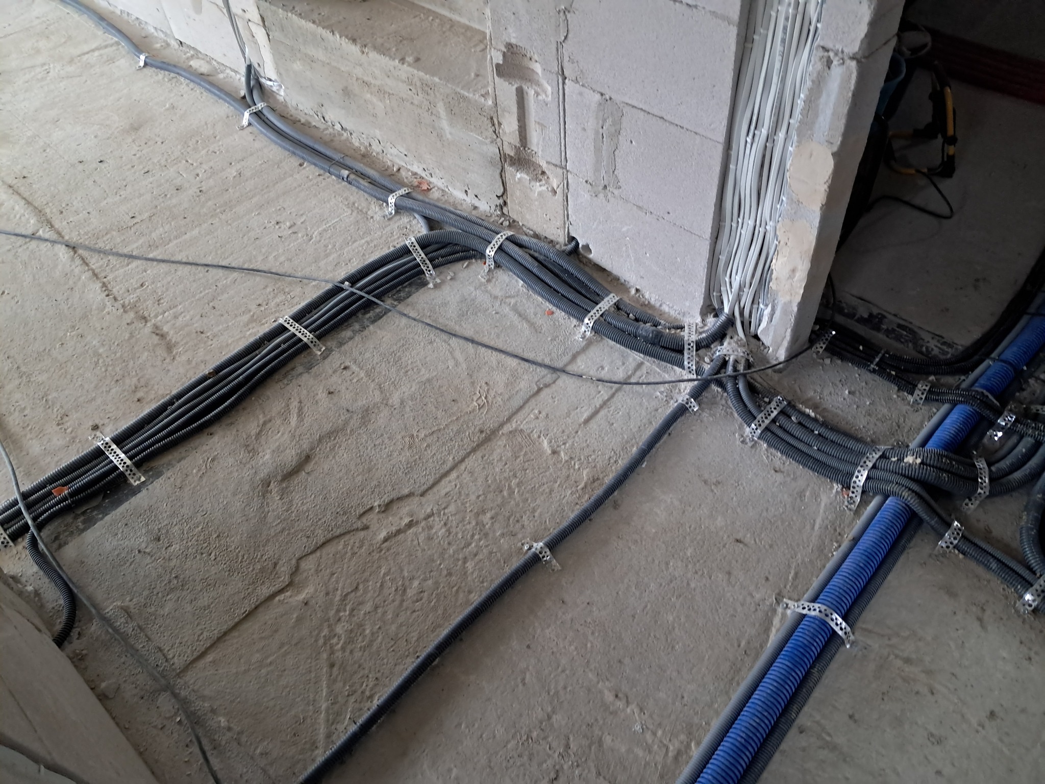 Szare i niebieskie kable w peszlach, przymocowane metalowymi uchwytami do surowej, betonowej podłogi i ściany z bloków. Widoczne białe przewody w ścianie.