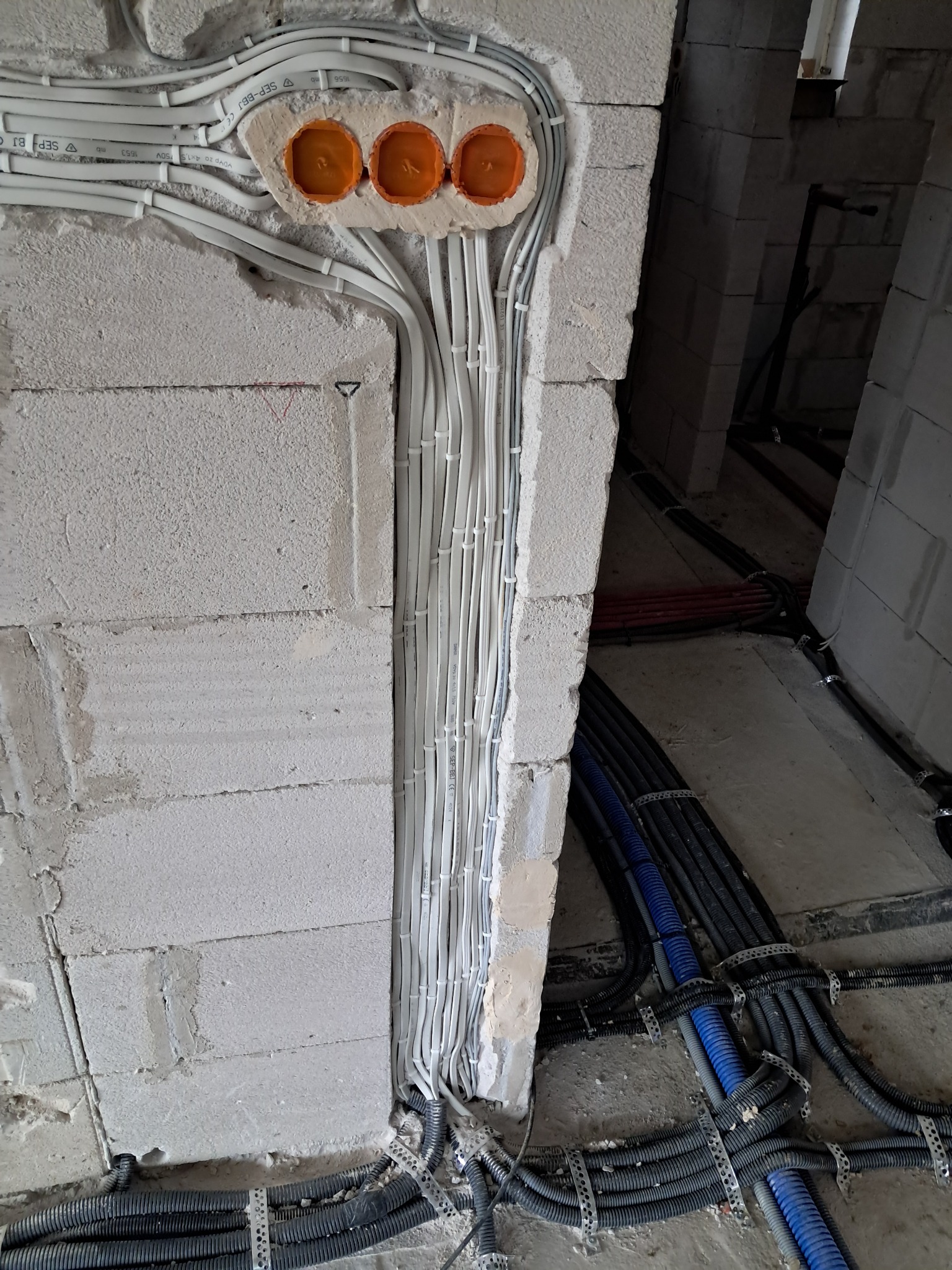 Instalacja elektryczna w trakcie montażu: białe kable prowadzone w ścianie z betonu komórkowego, pomarańczowe puszki pod gniazdka, rury karbowane na podłodze.