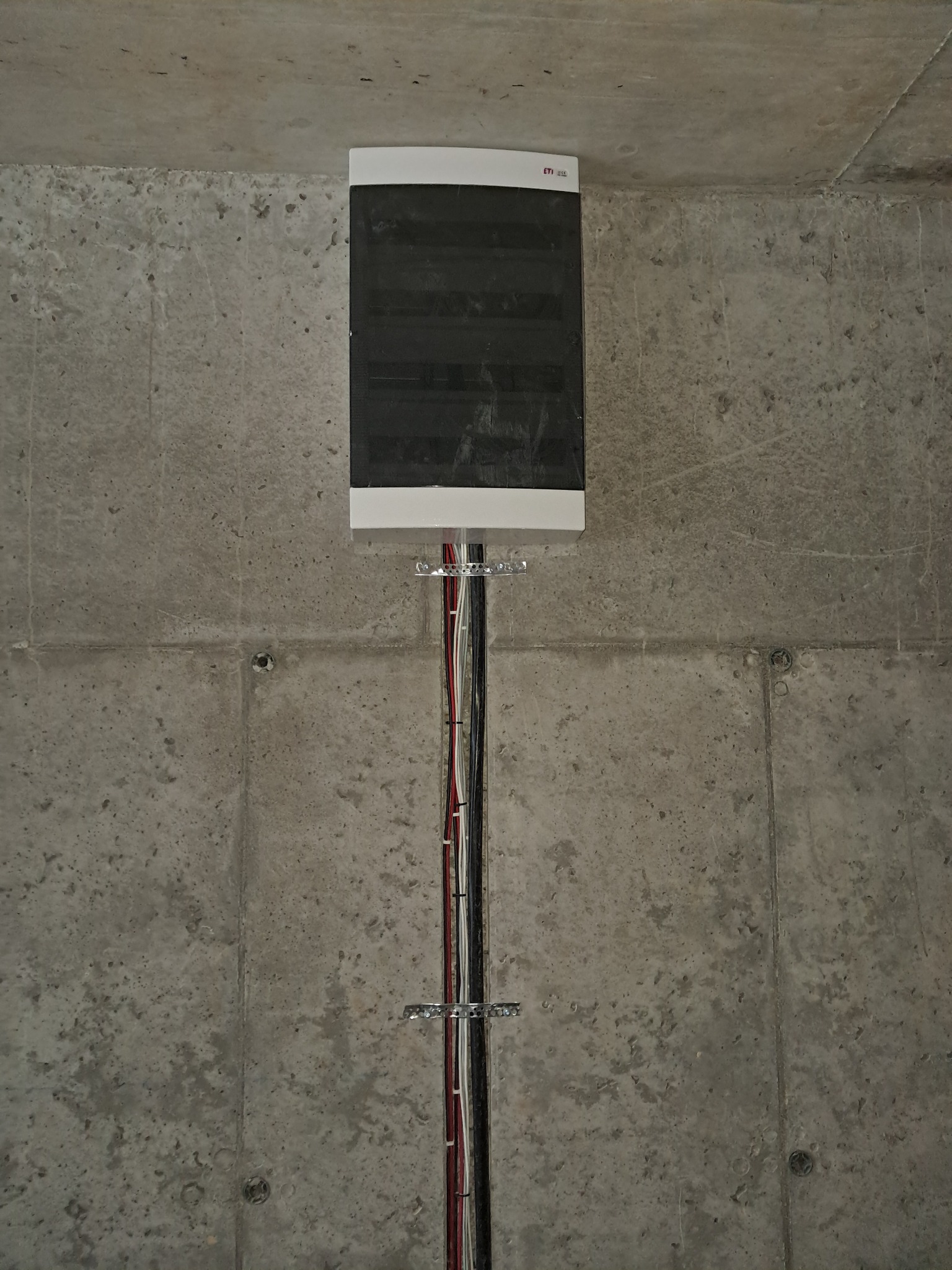 Zamknięta rozdzielnica elektryczna z przewodami przymocowanymi do surowego, betonowego stropu. Widoczne metalowe uchwyty mocujące.