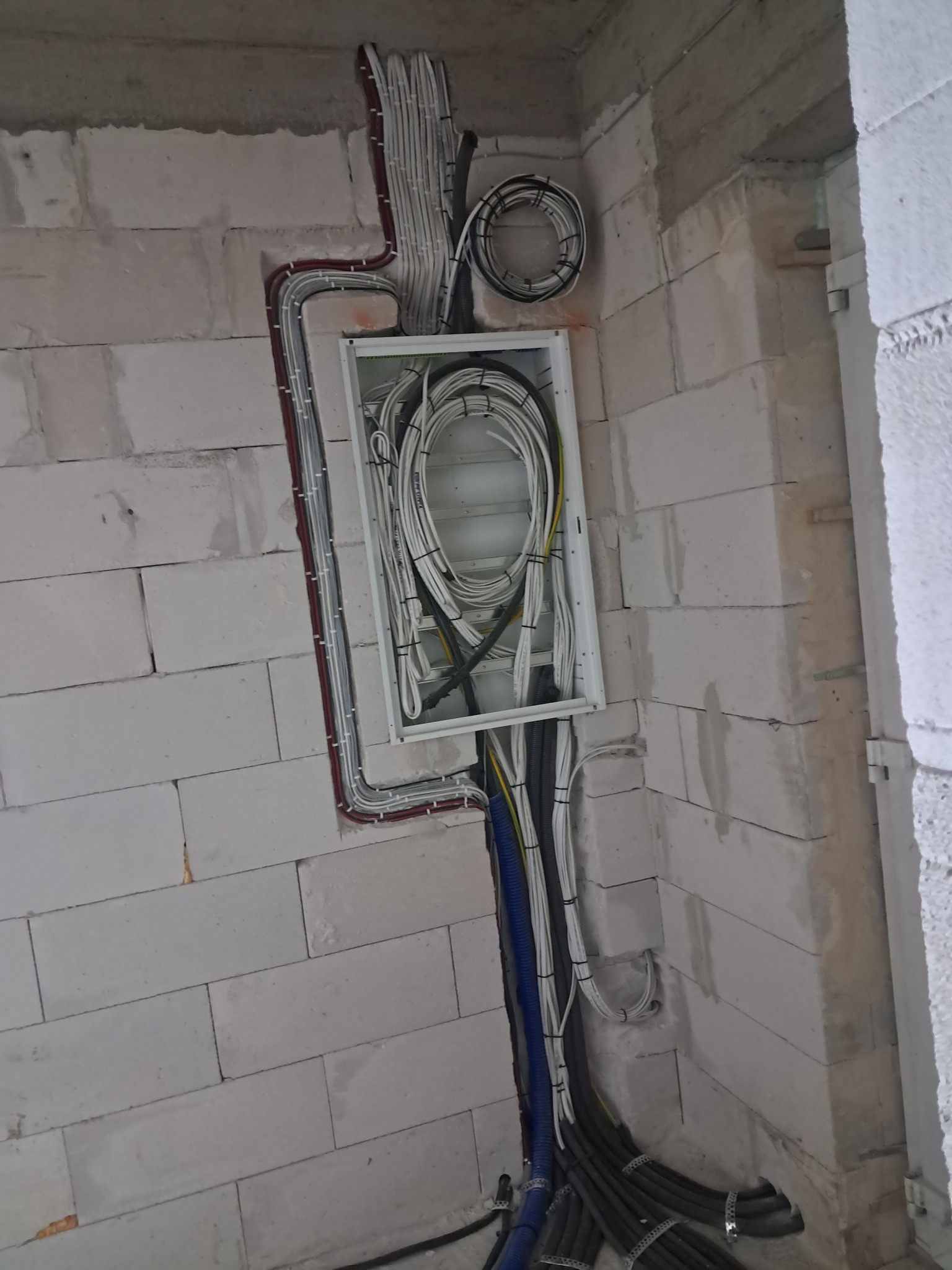 Rozdzielnia elektryczna z instalacją kablową w surowym wnętrzu budynku z białych bloczków. Kable prowadzone w peszlach i listwach, widoczne przewody w różnych kolorach.