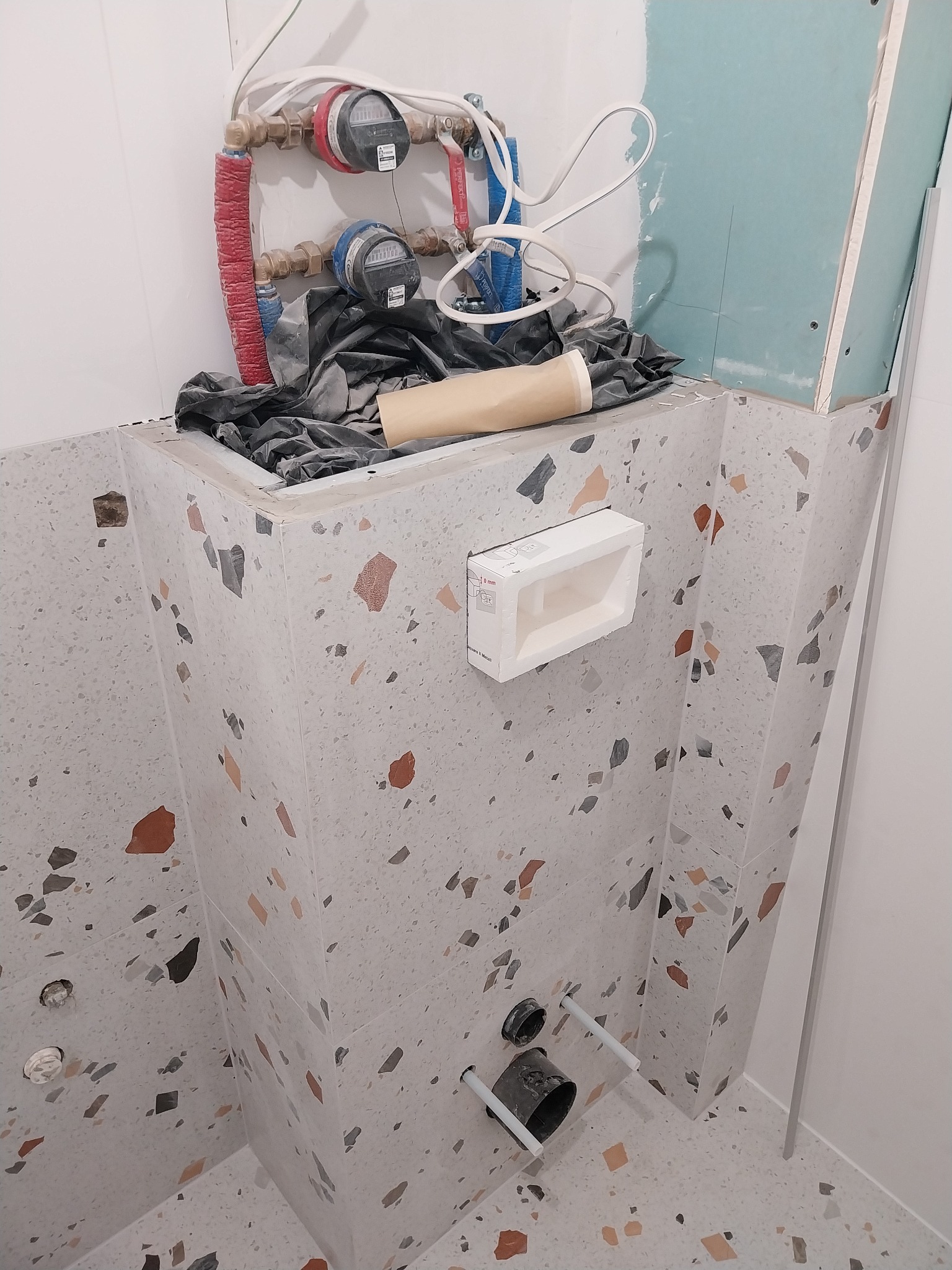 Narożnik łazienki w trakcie remontu z widoczną zabudową stelaża WC wykończoną płytkami lastryko, instalacją wodną z licznikami i fragmentem płyty kartonowo-gipsowej.
