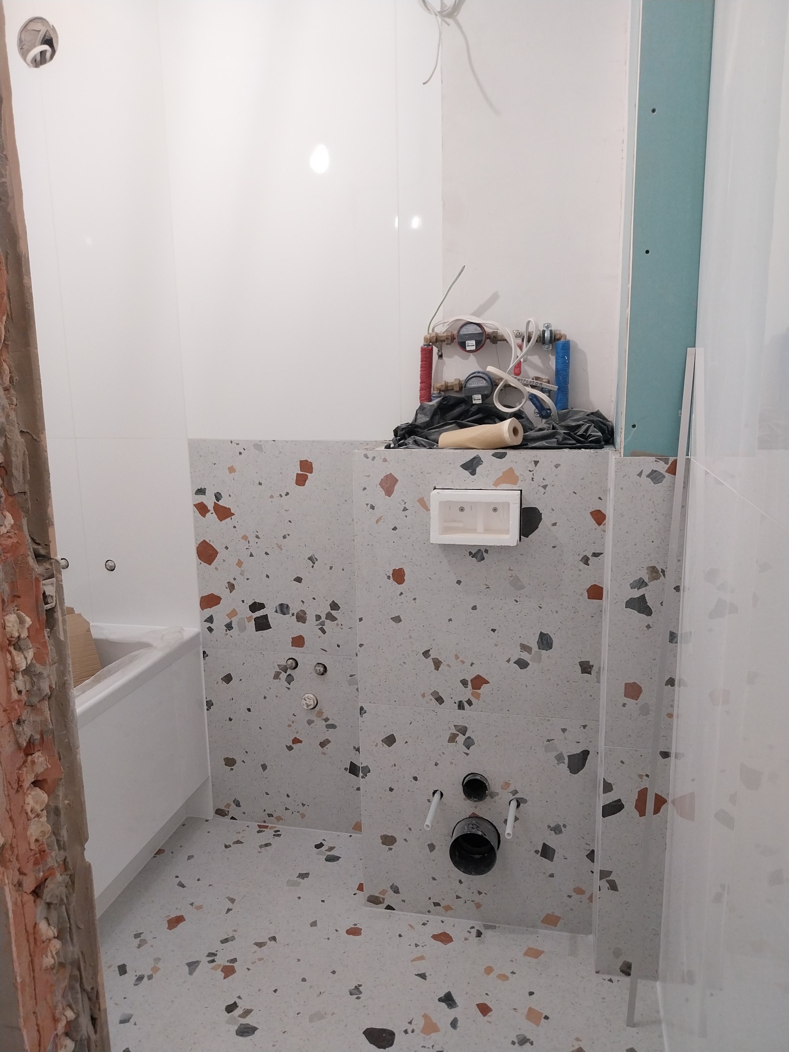 Łazienka w trakcie prac: białe płytki ścienne, terazzo na podłodze i obudowie stelaża WC, widoczne instalacje wodne, częściowo usunięta stara ściana.