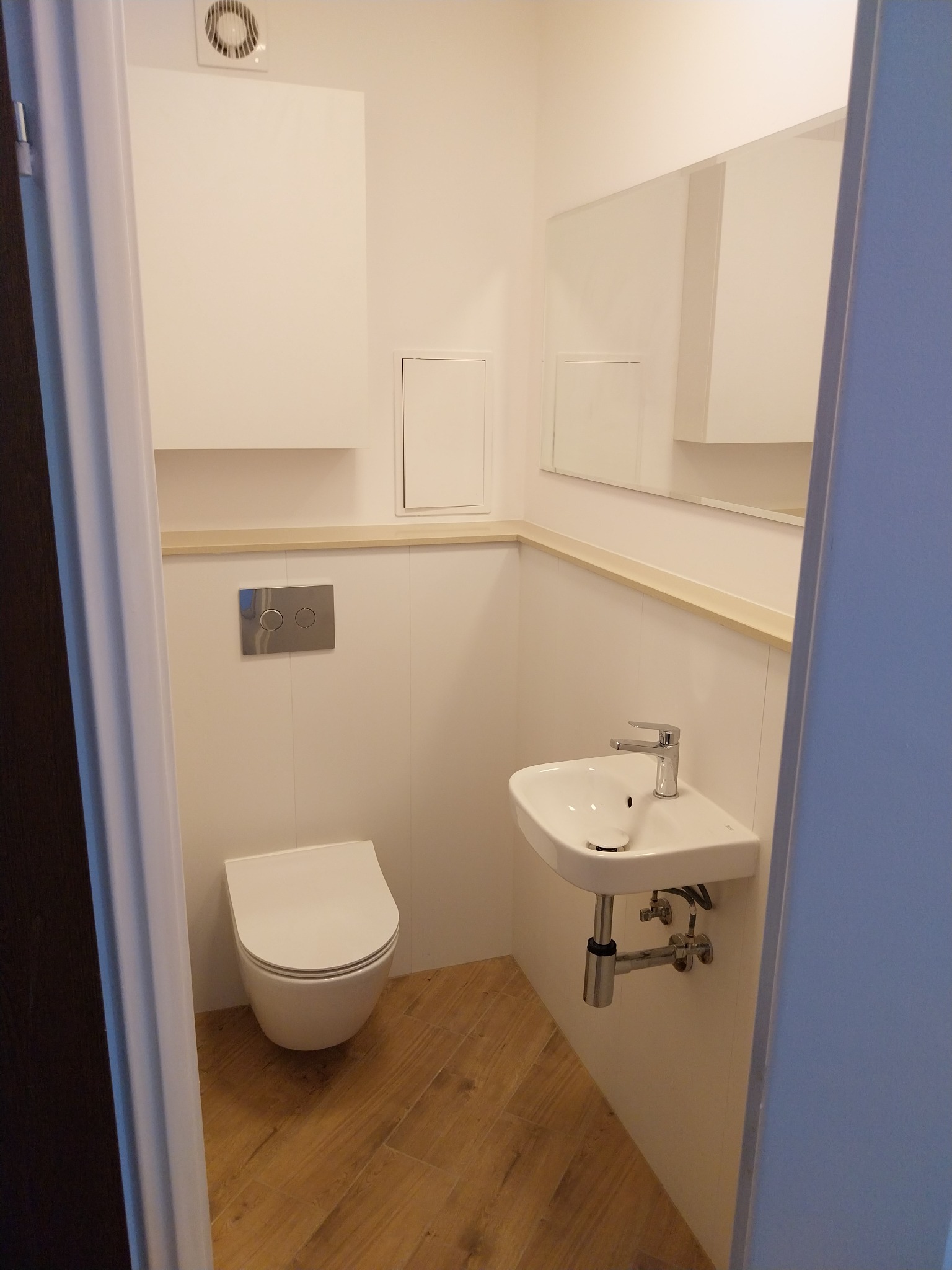 Nowoczesna, jasna toaleta z podwieszaną miską WC i umywalką. Drewnopodobne płytki podłogowe, minimalistyczny design, widok zza otwartych drzwi.