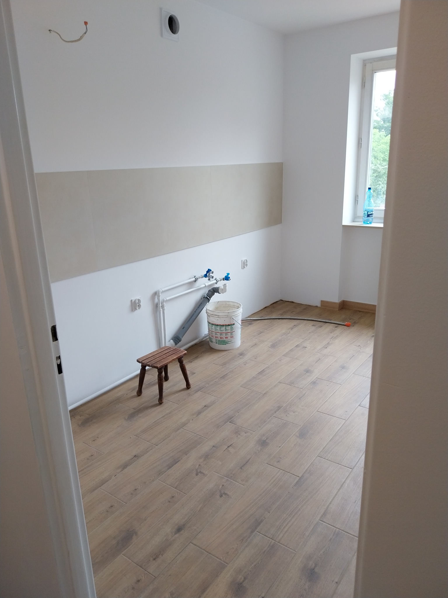 Łazienka w trakcie remontu: ułożona podłoga z płytek imitujących drewno, częściowo wykończone ściany z widoczną instalacją wodną i drobnym stołkiem.