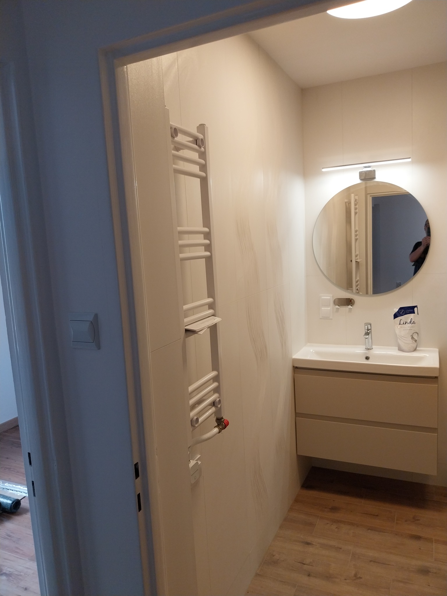 Jasna łazienka z grzejnikiem drabinkowym, okrągłym lustrem i szafką pod umywalką. Widoczne płytki imitujące drewno na podłodze i wzorzyste na ścianach.