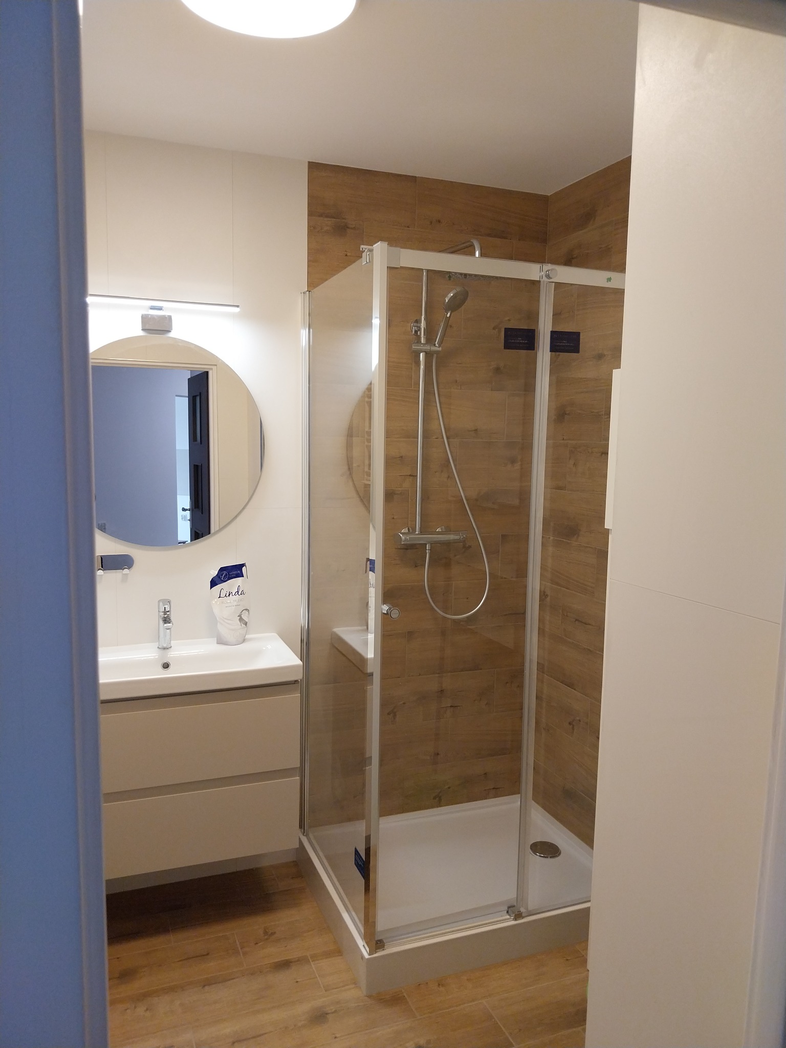 Nowoczesna łazienka po remoncie: kabina prysznicowa ze szklanymi drzwiami, okrągłe lustro nad umywalką z szafką, drewnopodobne płytki na ścianach i podłodze.