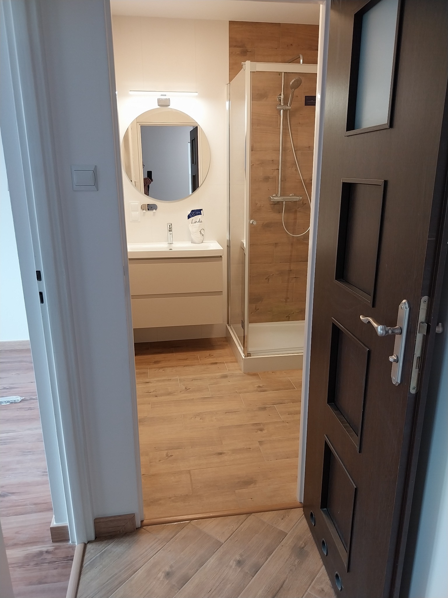 Nowoczesna łazienka z kabiną prysznicową, okrągłym lustrem i drewnopodobną podłogą, widziana zza uchylonych, ciemnych drzwi. Jasne, stonowane kolory.