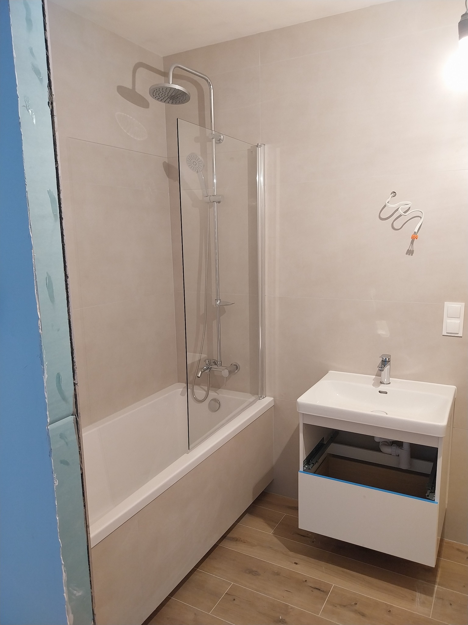 Wykończona łazienka z beżowymi płytkami, kabiną prysznicową nad wanną i białą szafką pod umywalką. Widoczne elementy instalacji elektrycznej.