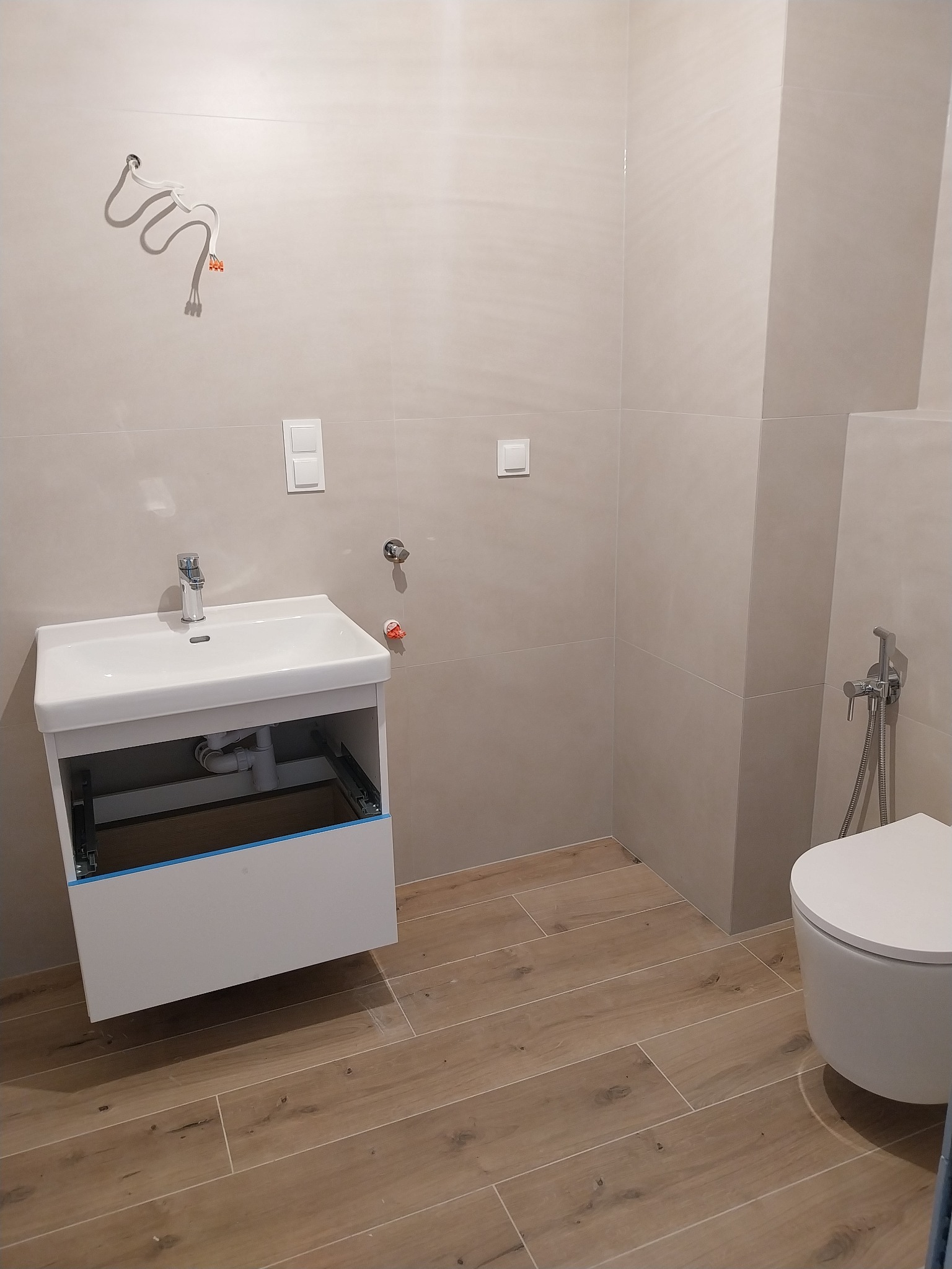 Jasna łazienka po remoncie: biała umywalka z szafką, wisząca toaleta z bidetką, płytki imitujące drewno na podłodze, beżowe płytki na ścianach. Widoczne przewody i instalacje.