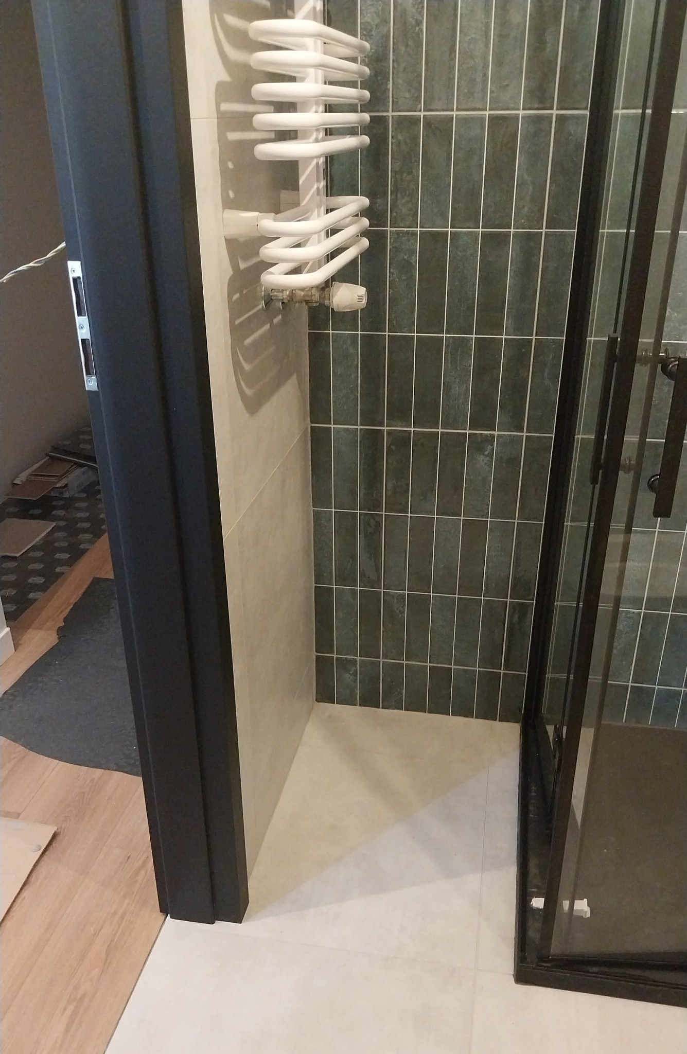 Wykończona łazienka z grzejnikiem drabinkowym, zielonymi płytkami i kabiną prysznicową z czarnymi profilami. Widok zza czarnej ościeżnicy.