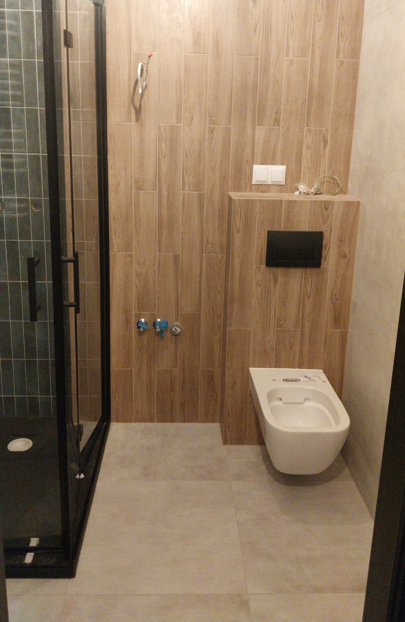 Nowoczesna łazienka z kabiną prysznicową typu walk-in, drewnopodobnymi płytkami na ścianie i podwieszaną toaletą. Widoczne niedokończone prace instalacyjne.