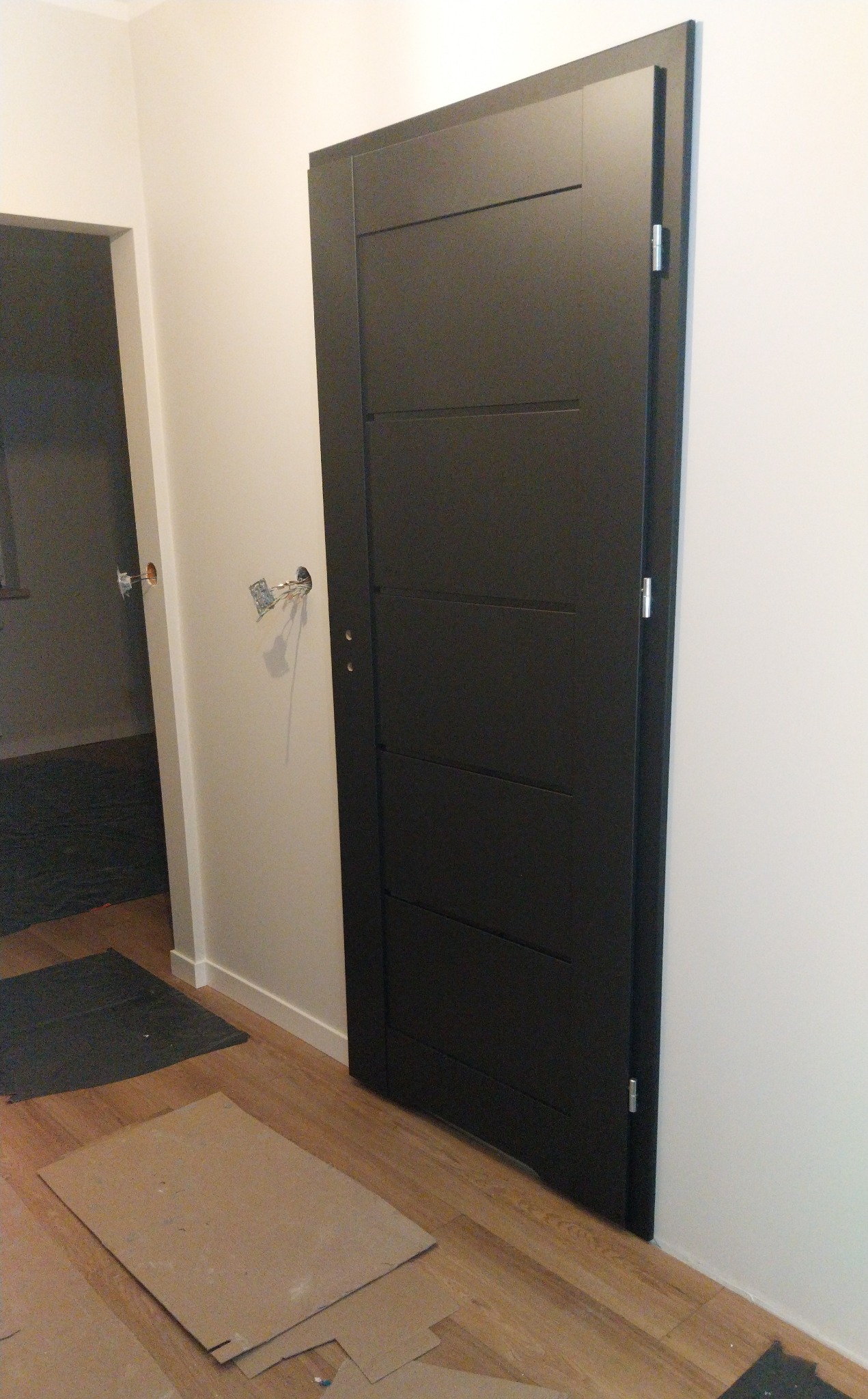 Ciemne, nowoczesne drzwi wewnętrzne z trzema poziomymi wgłębieniami, zamontowane w jasnym pomieszczeniu z drewnianą podłogą. Widoczne przygotowania do prac wykończeniowych.