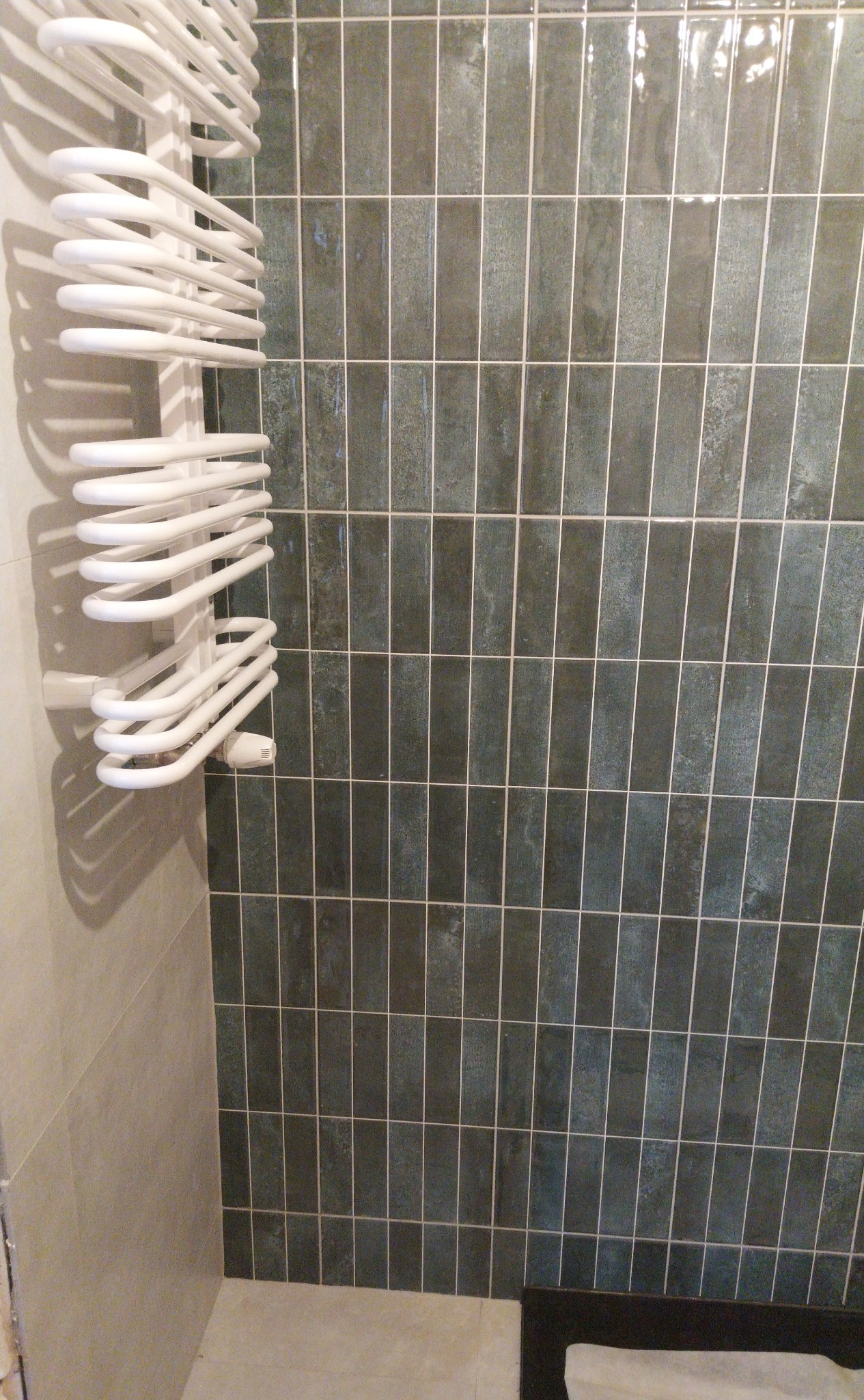 Błyszczące, pionowe, zielono-szare płytki w łazience z białym grzejnikiem drabinkowym po lewej stronie. Widoczny fragment podłogi i ściany w jasnym odcieniu.