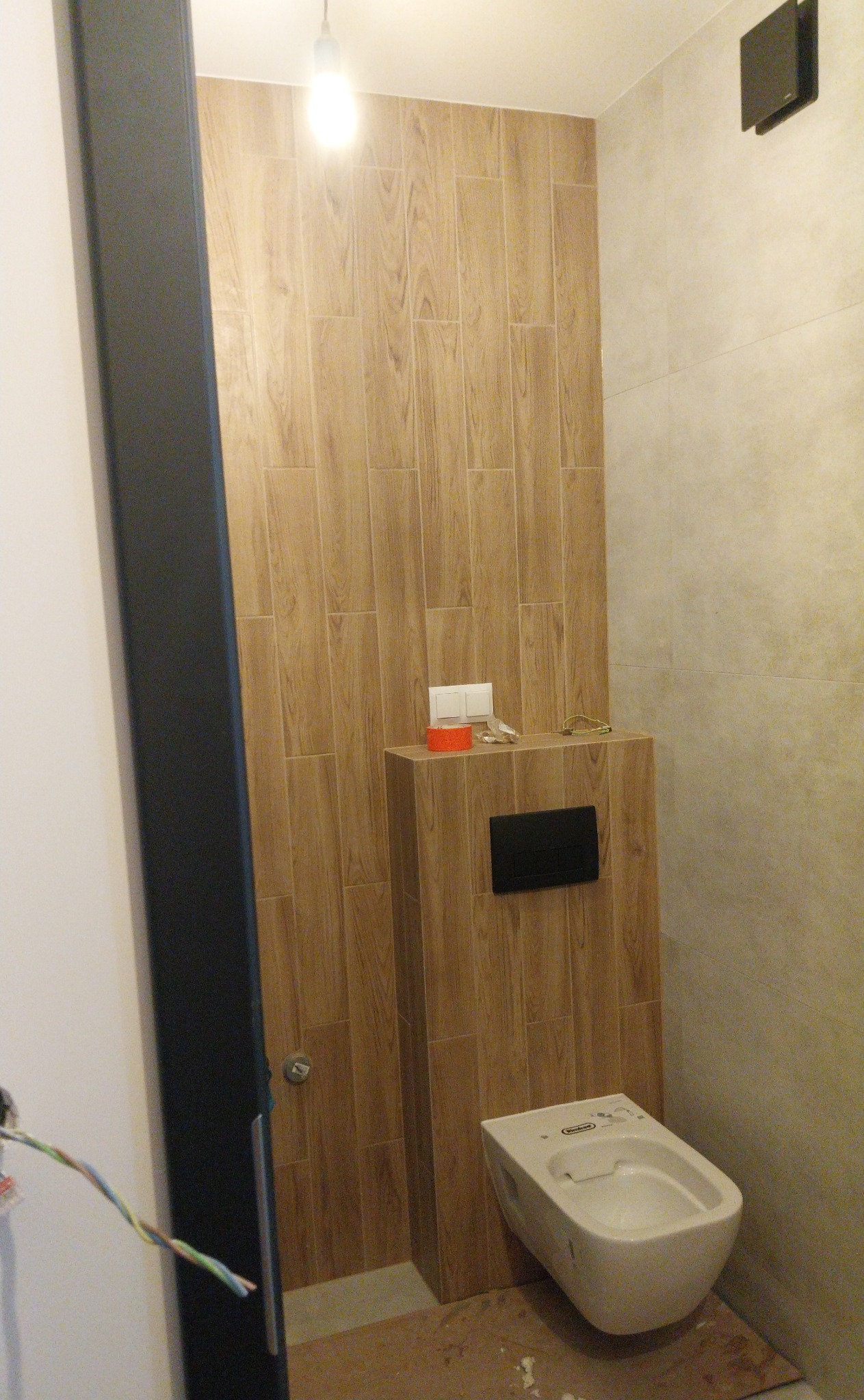 Nowoczesna łazienka w trakcie wykończenia: drewnopodobne płytki na ścianie, podwieszana toaleta, widoczne przewody elektryczne.