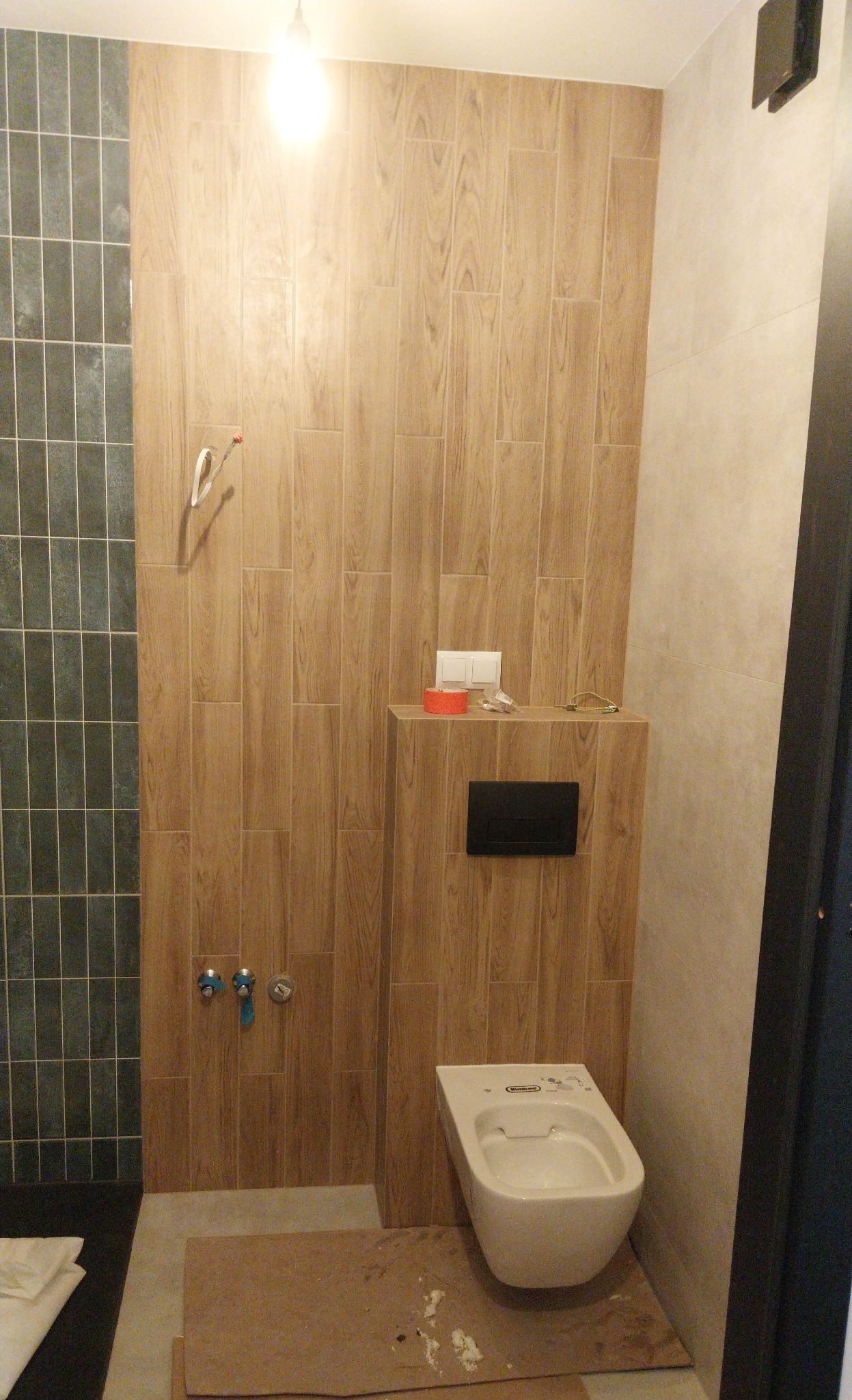 Wykończona łazienka z drewnopodobnymi płytkami na ścianie, wiszącą toaletą i zielonymi płytkami w strefie prysznica. Widoczne elementy instalacji i akcesoria.