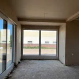 Wnętrze w stanie deweloperskim po nałożeniu jasnobeżowych tynków, widoczne duże okna z widokiem na zewnątrz, surowy betonowy strop i wystające kable elektryczne.