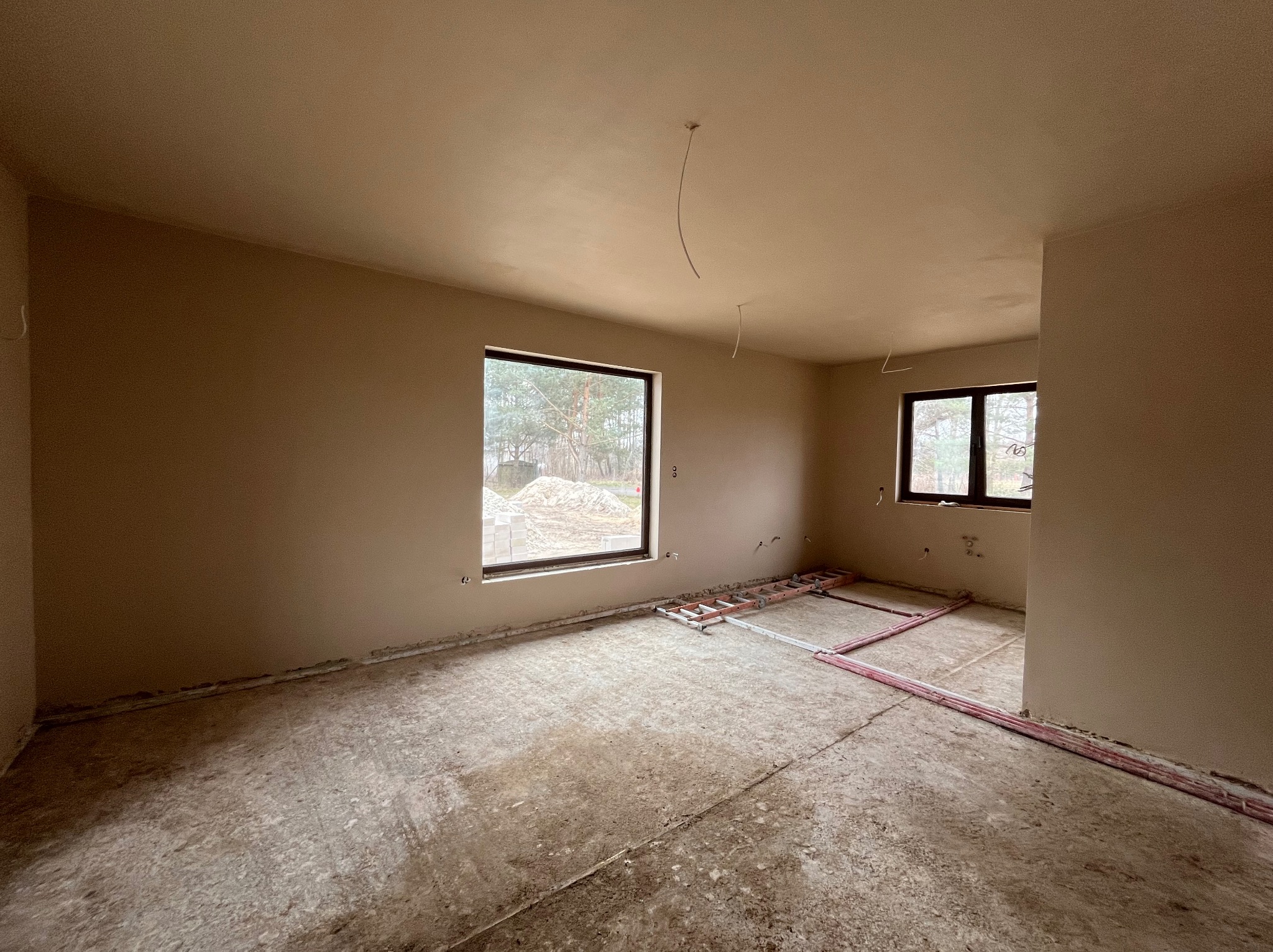 Pokój w trakcie prac wykończeniowych: ściany i sufit pokryte świeżym, jasnym tynkiem, widoczne kable zwisające z sufitu, na podłodze rozprowadzone rurki ogrzewania podłogowego i drabina. Za oknem...