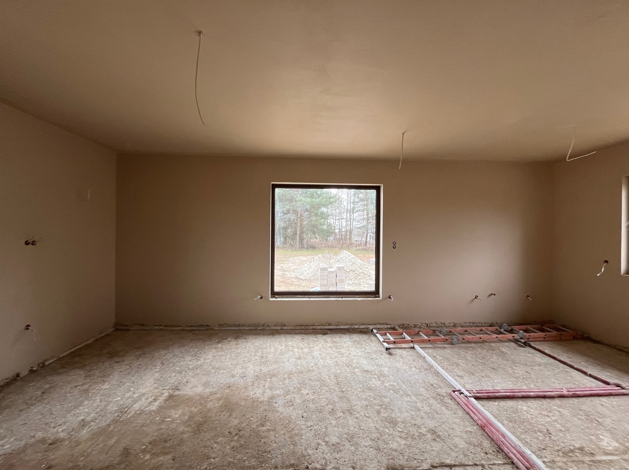 Wnętrze pokoju po nałożeniu jasnego tynku maszynowego, widoczne wystające przewody elektryczne, grzejniki podłogowe oraz drabina na podłodze.