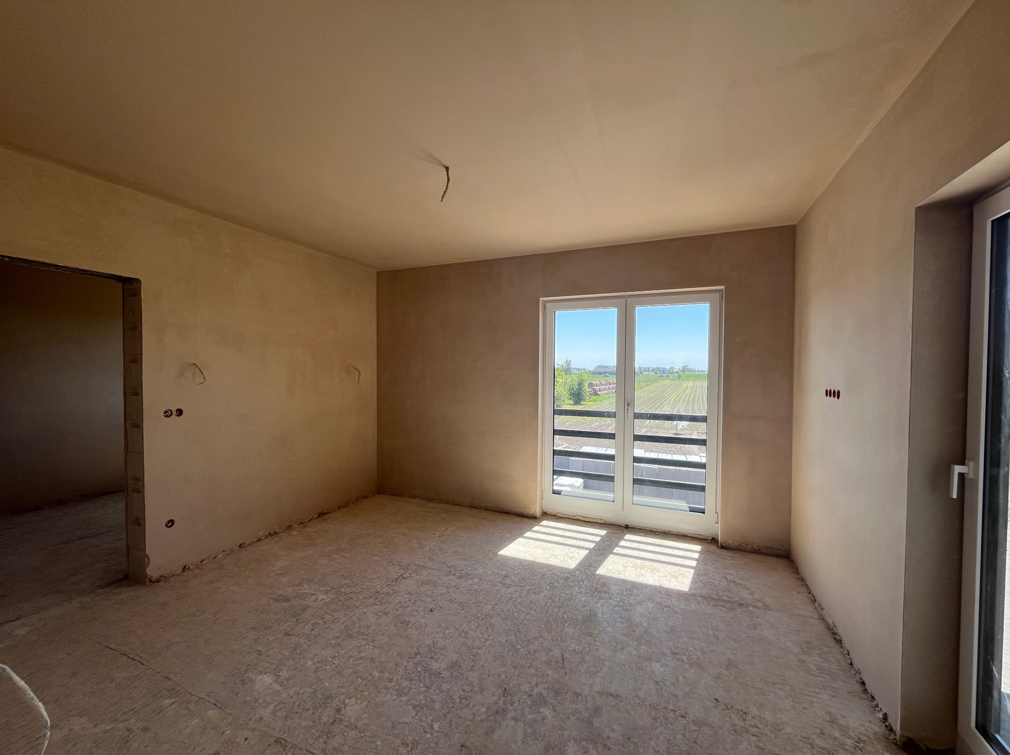 Wnętrze surowego pomieszczenia z nałożonymi jasnymi tynkami, widok na pole przez okno balkonowe, widoczne wyprowadzenia instalacji elektrycznej.