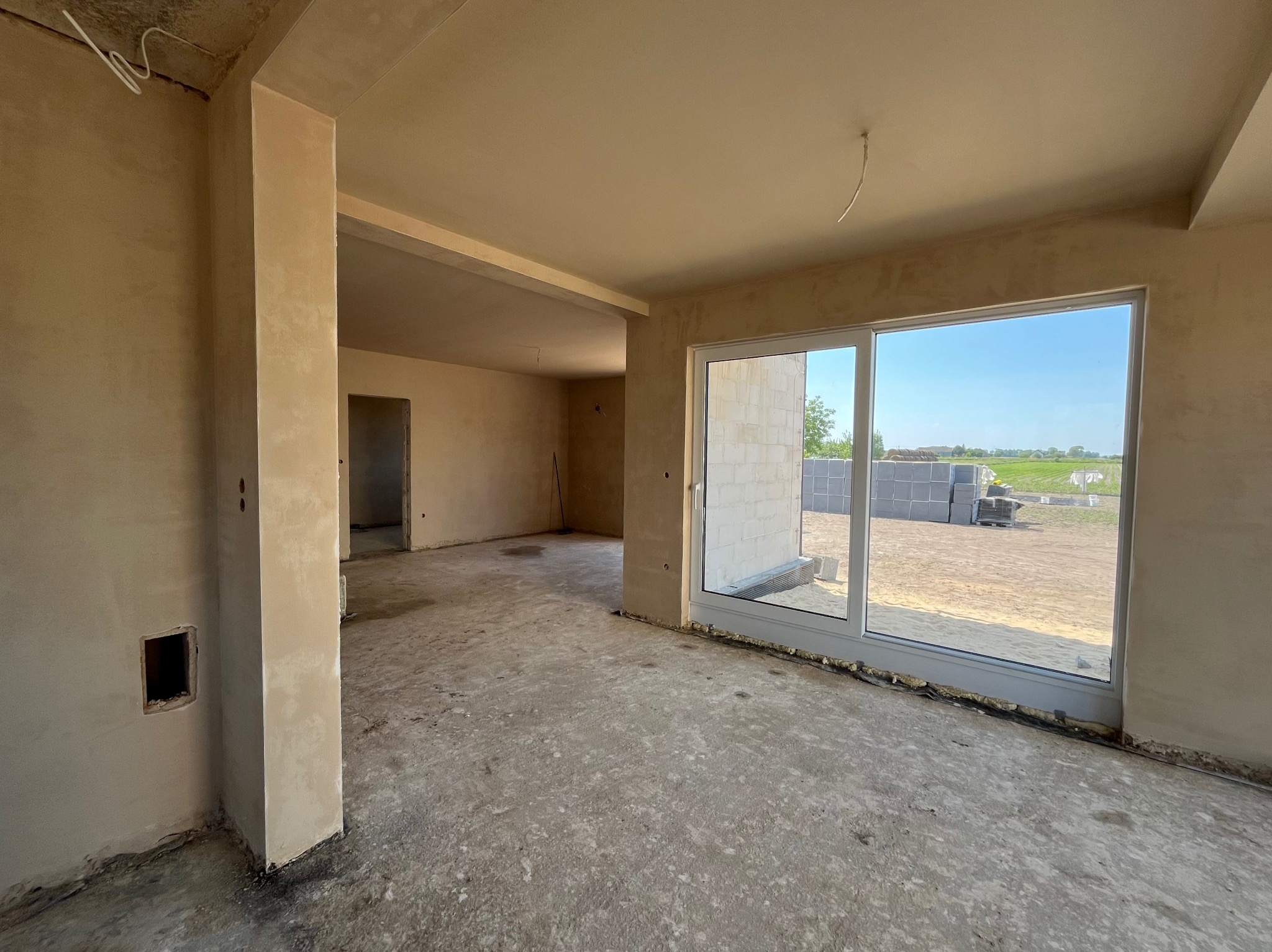Surowe wnętrze domu jednorodzinnego po tynkowaniu maszynowym; widoczne ściany i sufit pokryte świeżym tynkiem, betonowa podłoga oraz okno z widokiem na plac budowy z ułożonymi bloczkami.