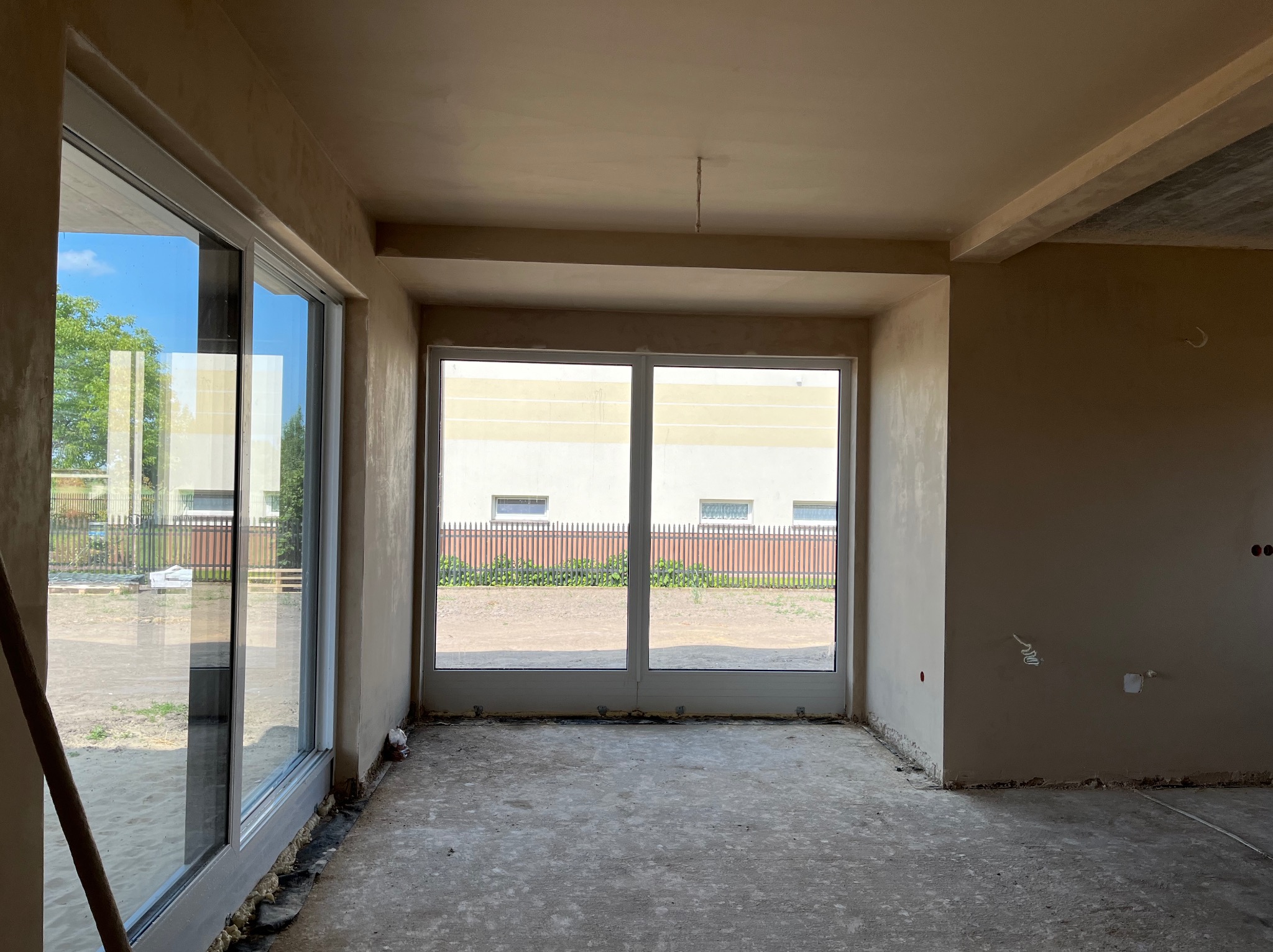 Wnętrze w stanie deweloperskim po nałożeniu jasnobeżowych tynków, widoczne duże okna z widokiem na zewnątrz, surowy betonowy strop i wystające kable elektryczne.