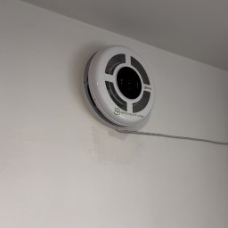 Eryk Suder Ecoventure - Okrągły, biały anemostat z kratką i logo 'ECOVENTURE' zamontowany na białej ścianie, z widocznym szarym przewodem.