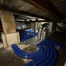 Eryk Suder Ecoventure - Niebieskie rury rekuperacji rozchodzące się z metalowej skrzynki rozdzielczej w przestrzeni poddasza z drewnianym stropem, widoczne przewody elektryczne i fragment ściany z cegieł.