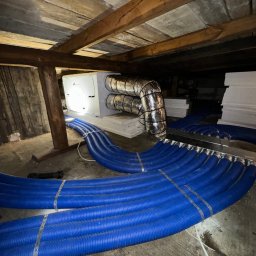 Eryk Suder Ecoventure - Widok instalacji rekuperacji z niebieskimi przewodami wentylacyjnymi w surowym pomieszczeniu poddasza z drewnianą konstrukcją stropu.