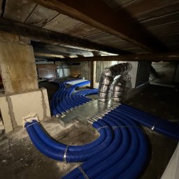 Eryk Suder Ecoventure - Instalacja przewodów wentylacyjnych w przestrzeni poddasza, widoczne niebieskie rury karbowane z metalowymi obejmami, srebrne przewody izolowane oraz elementy konstrukcyjne dachu.