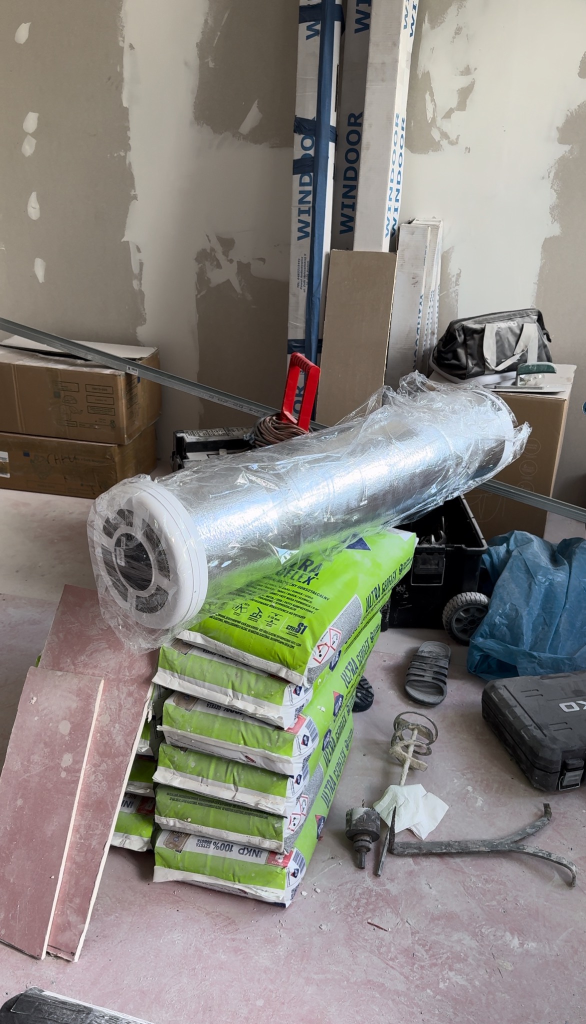 Przygotowanie do montażu systemu wentylacyjnego: izolowany przewód wentylacyjny w folii, stos worków z zaprawą budowlaną, narzędzia i materiały budowlane w surowym wnętrzu, widoczne profile i płyty...