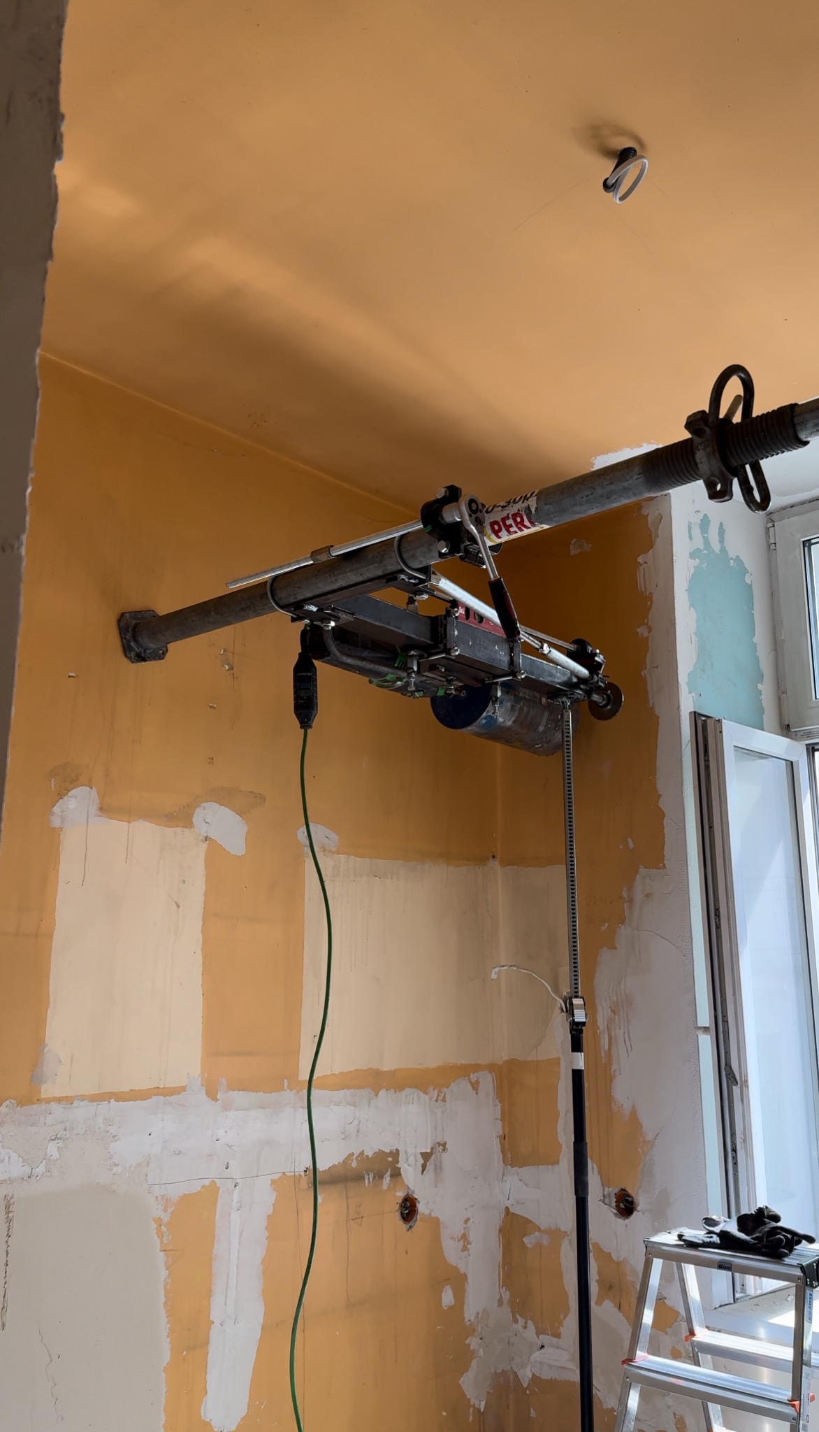 Montaż instalacji wentylacyjnej z użyciem specjalistycznego podnośnika sufitowego PERI na tle przygotowywanej ściany z widocznymi śladami po kuciu i szpachlowaniu.