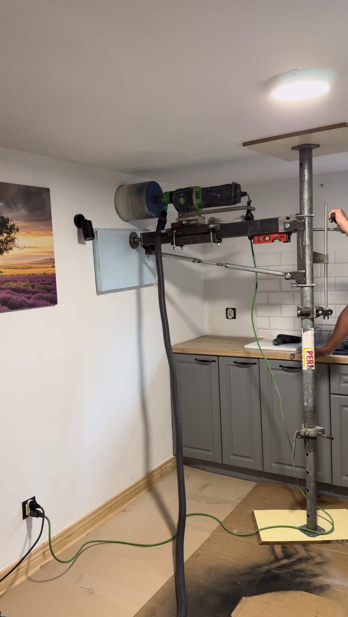Wiertnica diamentowa mocowana do ściany za pomocą specjalnego stelaża w trakcie wiercenia otworu w pomieszczeniu z szarymi szafkami kuchennymi, widoczny przewód odkurzacza odprowadzający urobek.