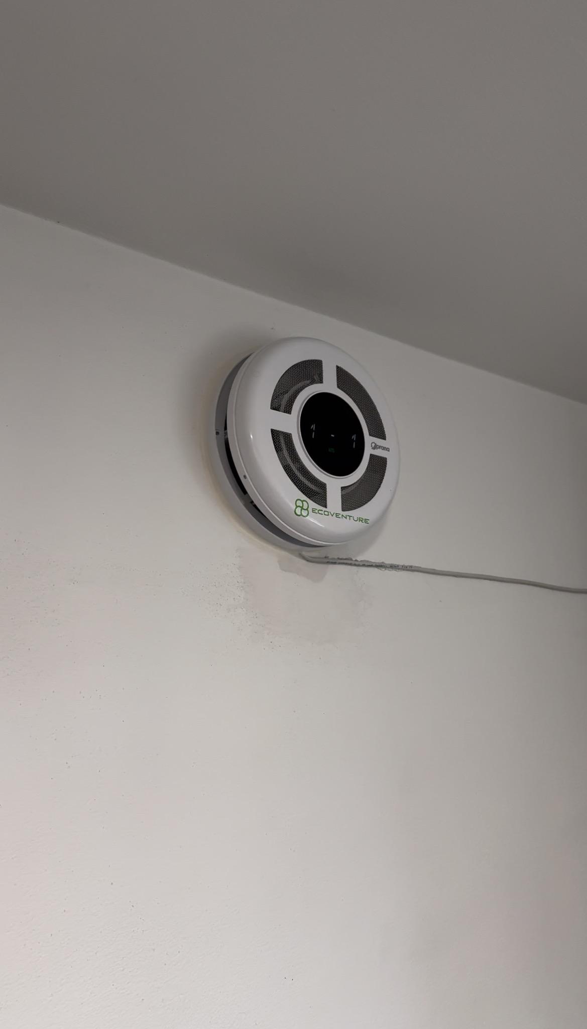 Okrągły, biały anemostat z kratką i logo 'ECOVENTURE' zamontowany na białej ścianie, z widocznym szarym przewodem.