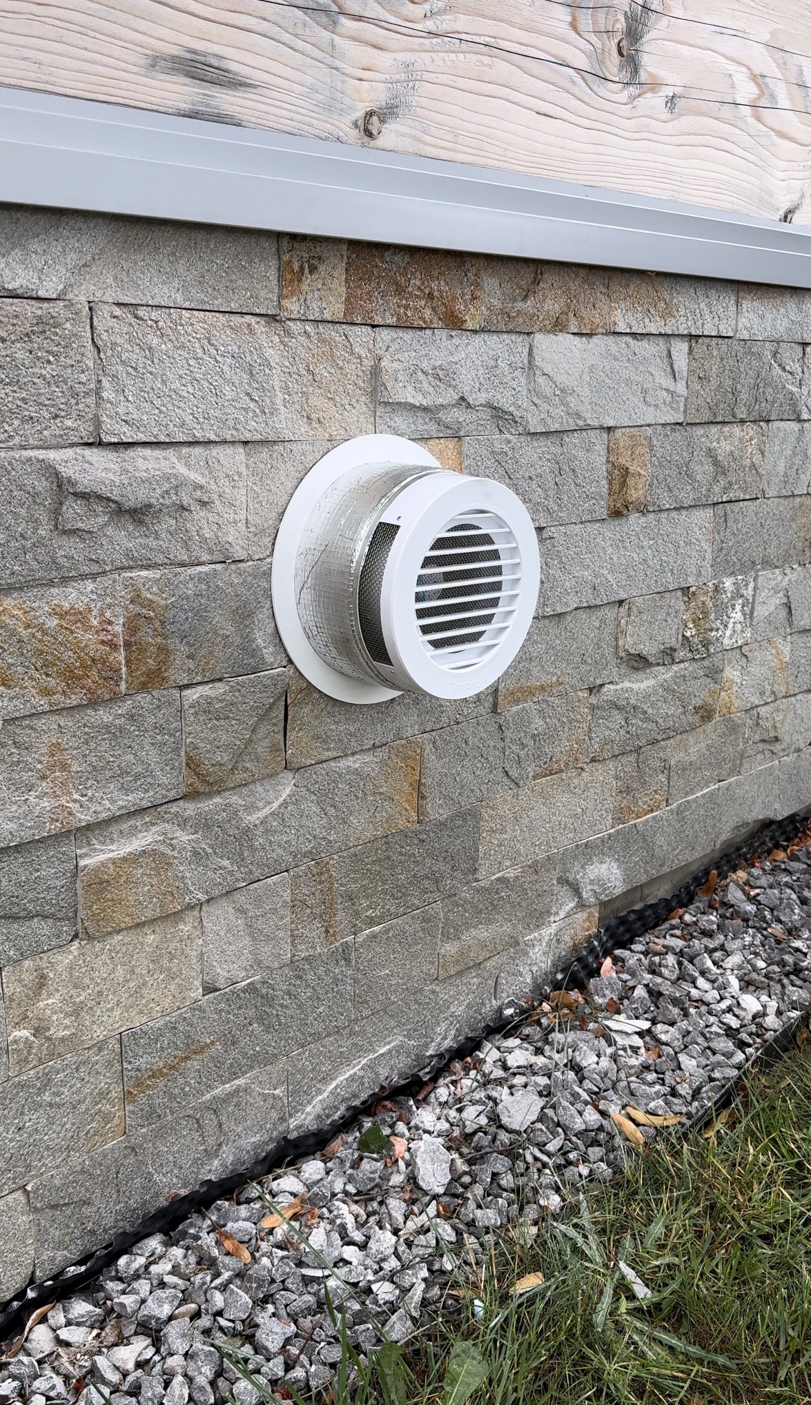 Biała, okrągła kratka wentylacyjna zamontowana na elewacji z szarego kamienia, widoczna aluminiowa rura wentylacyjna, u dołu kamienny drenaż.