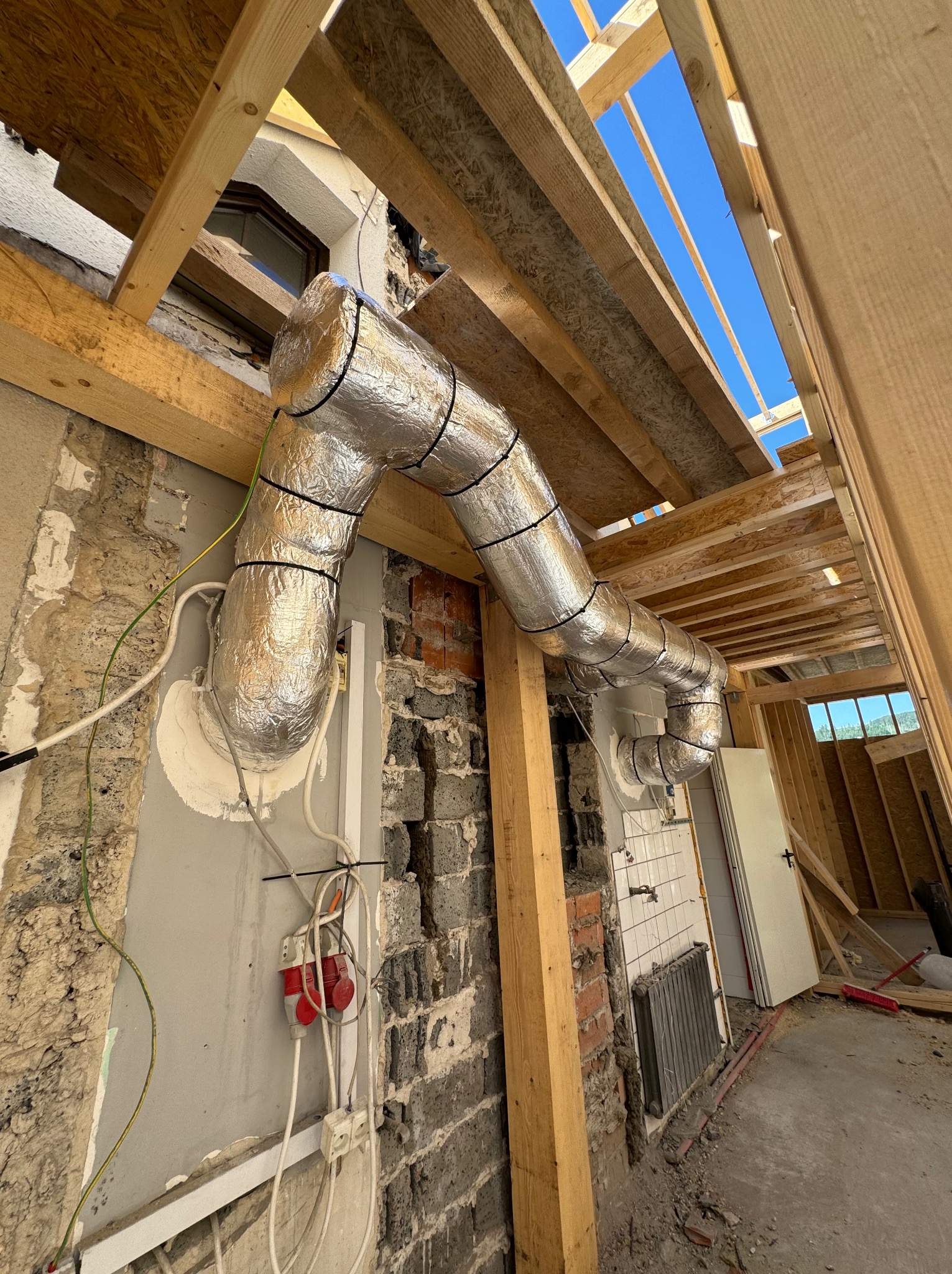 Instalacja przewodów wentylacyjnych w izolacji aluminiowej w surowym wnętrzu budynku w trakcie przebudowy, widoczne elementy konstrukcji drewnianej dachu i fragmenty instalacji elektrycznej.