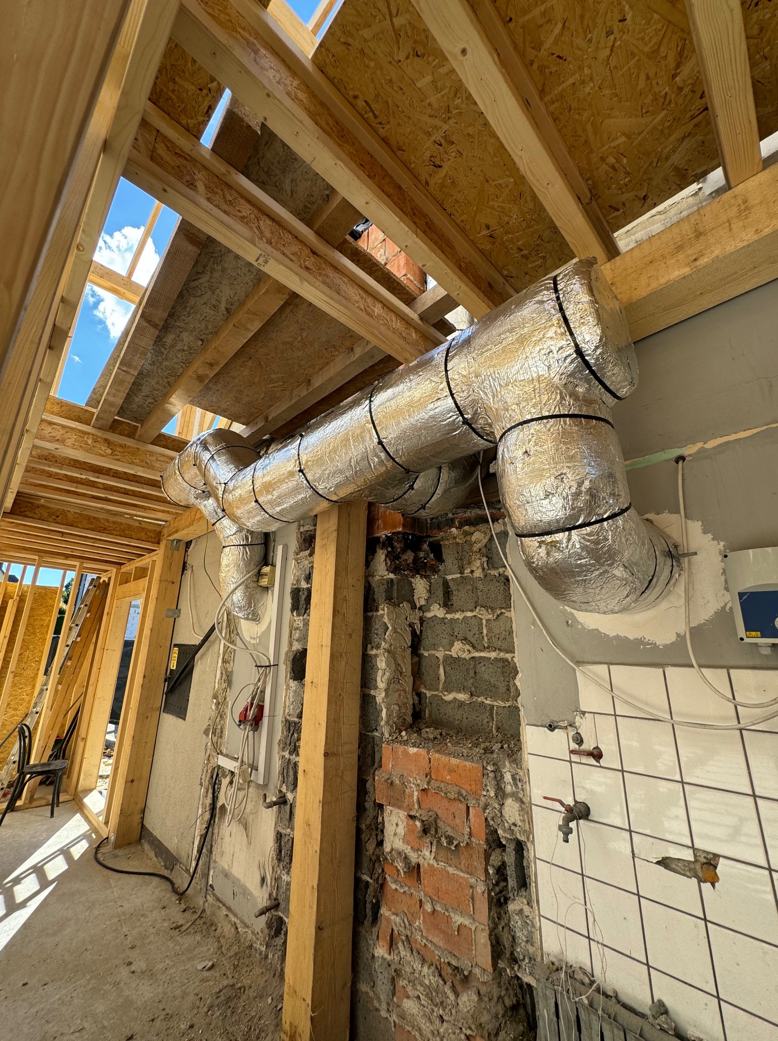 Wnętrze w trakcie remontu z widocznymi, zaizolowanymi przewodami wentylacyjnymi pod sufitem z drewnianą konstrukcją, fragment ściany z cegły i instalacją wodną.