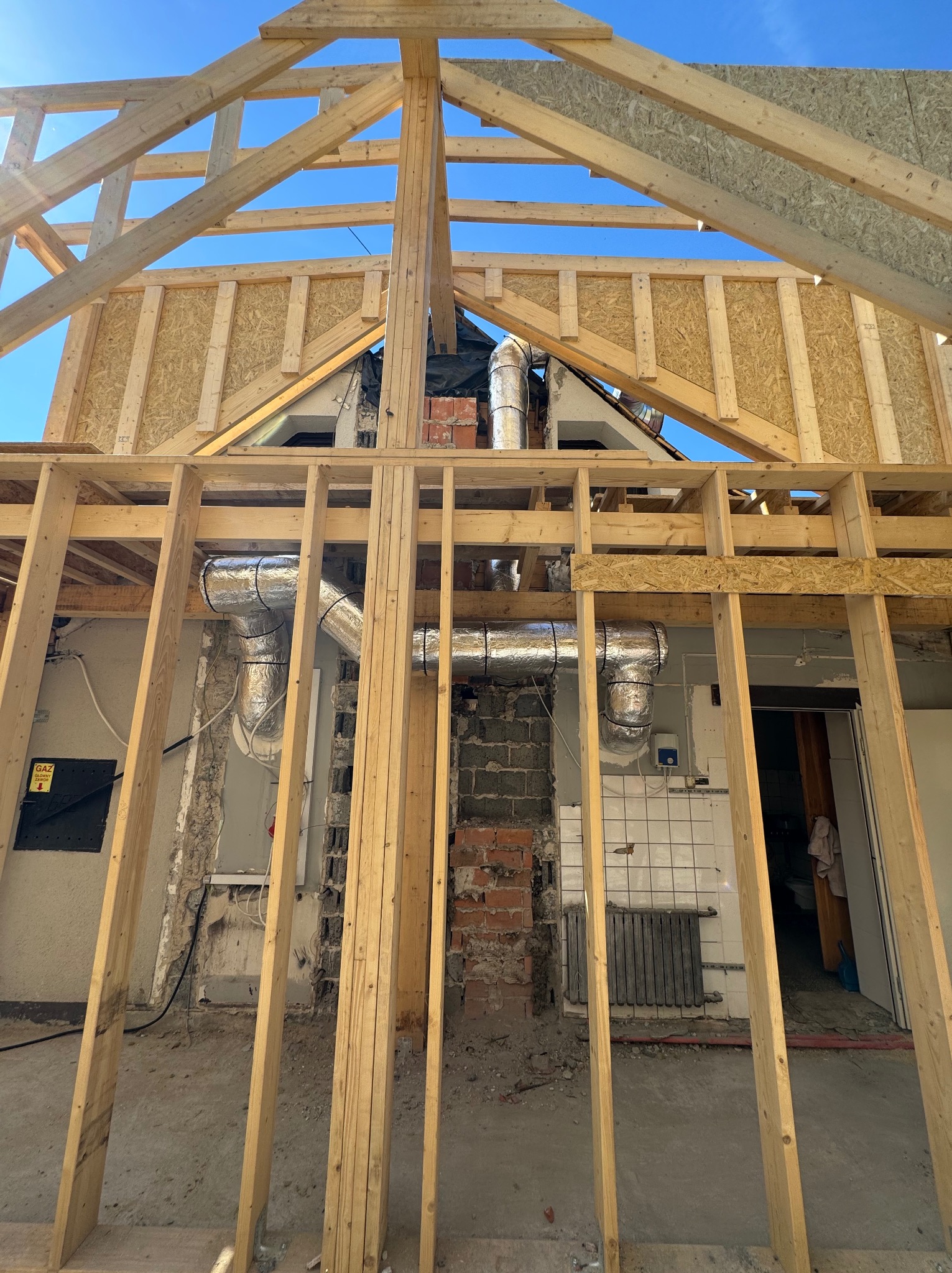 Wnętrze budynku w trakcie montażu instalacji wentylacyjnej, widoczne aluminiowe rury wentylacyjne, drewniana konstrukcja szkieletowa dachu i ścian, oraz fragmenty starej instalacji grzewczej...