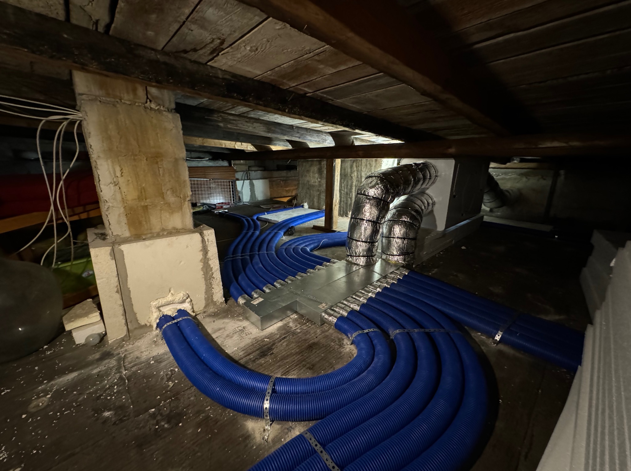 Niebieskie rury rekuperacji rozchodzące się z metalowej skrzynki rozdzielczej w przestrzeni poddasza z drewnianym stropem, widoczne przewody elektryczne i fragment ściany z cegieł.