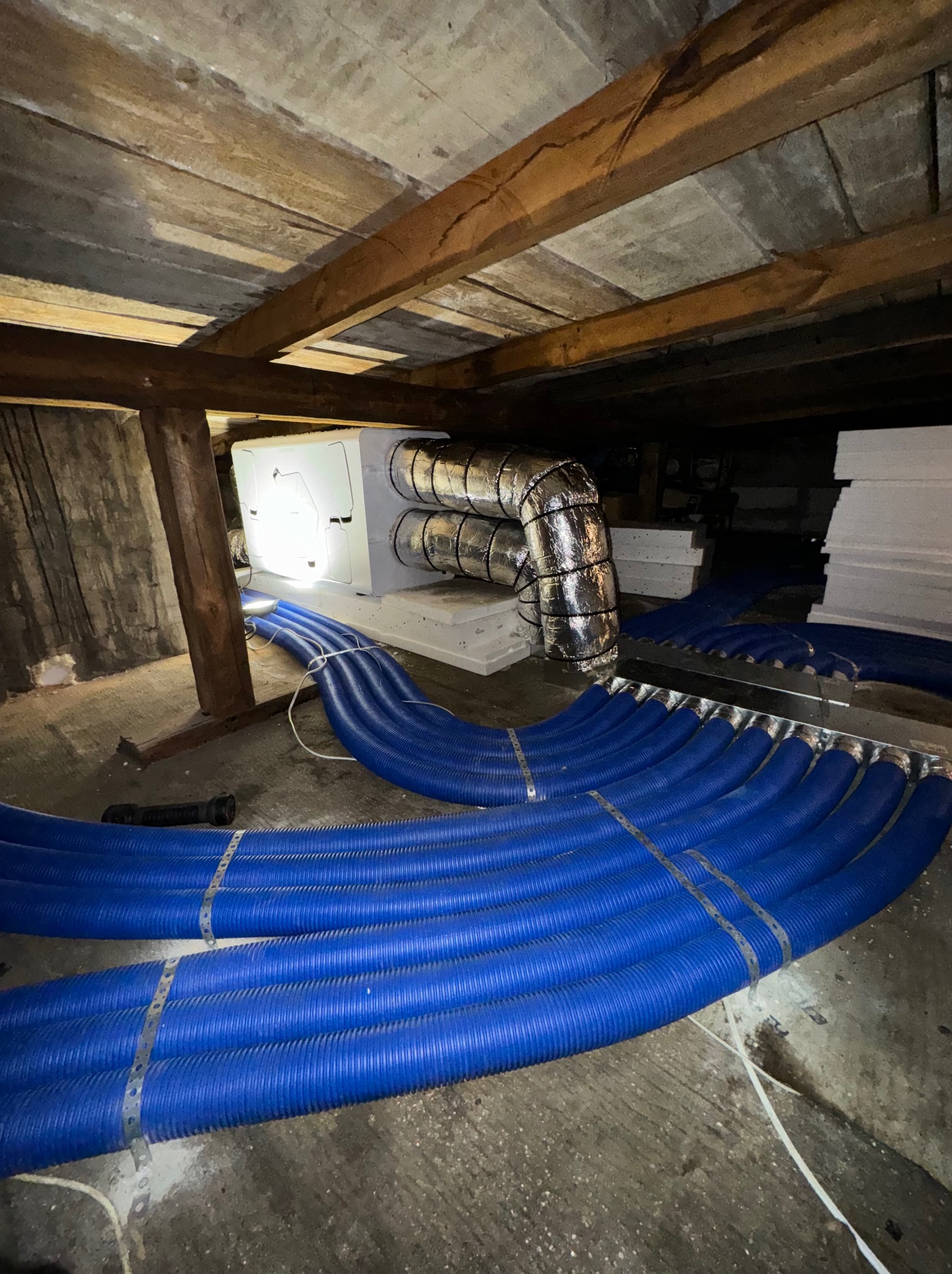 Widok instalacji rekuperacji z niebieskimi przewodami wentylacyjnymi w surowym pomieszczeniu poddasza z drewnianą konstrukcją stropu.