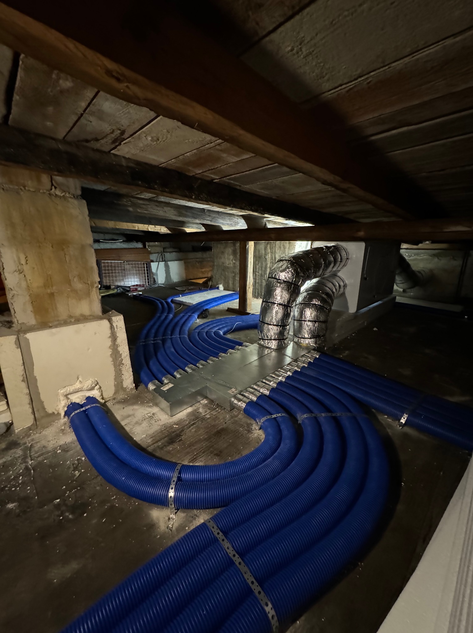 Instalacja przewodów wentylacyjnych w przestrzeni poddasza, widoczne niebieskie rury karbowane z metalowymi obejmami, srebrne przewody izolowane oraz elementy konstrukcyjne dachu.