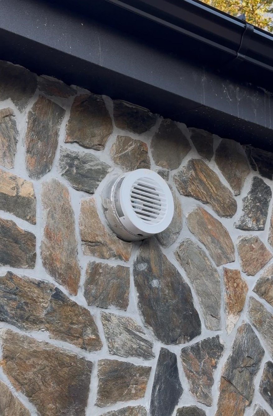 Biała okrągła kratka wentylacyjna zamontowana na elewacji z nieregularnych kamieni w odcieniach brązu i szarości, widoczny fragment czarnej rynny dachowej