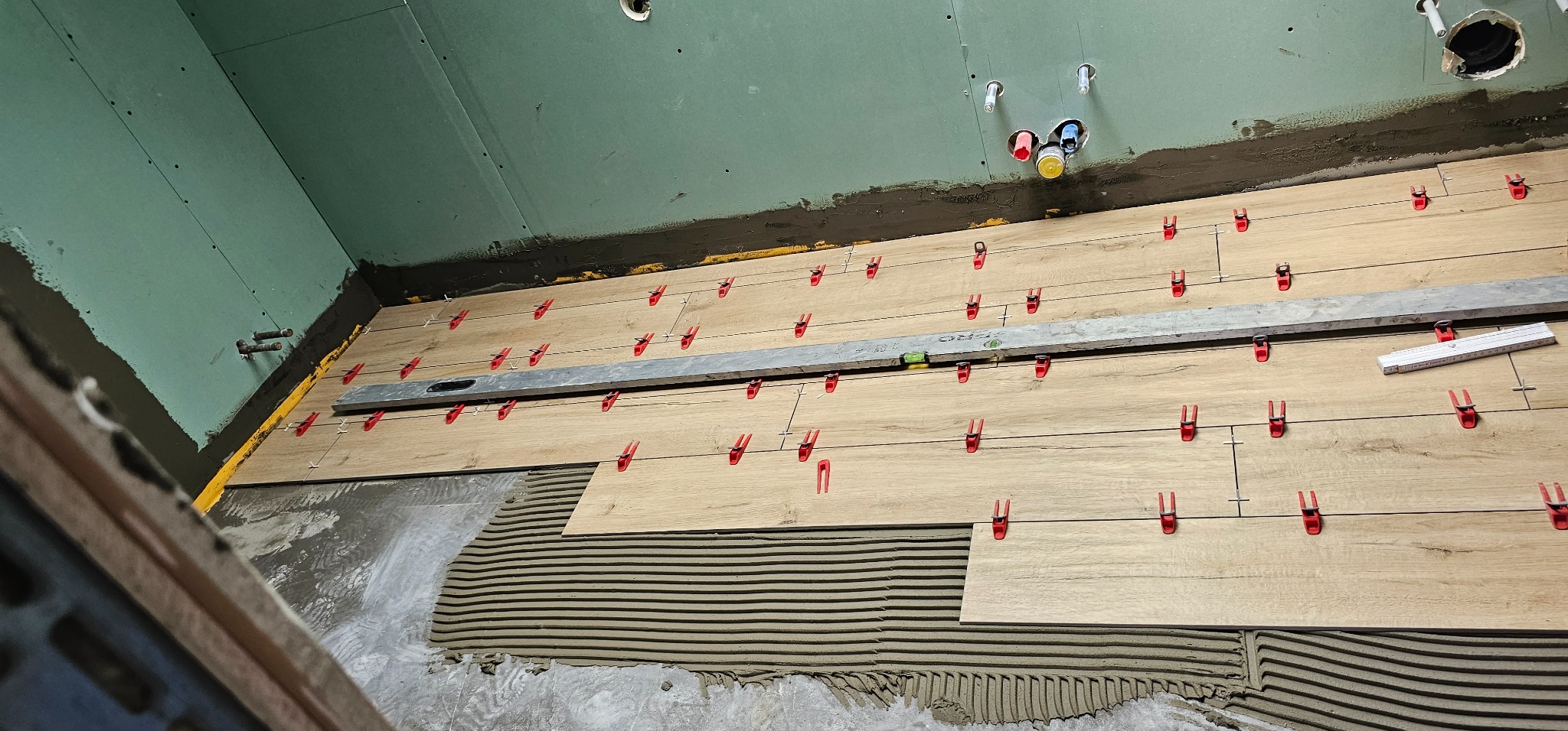 Układanie płytek imitujących drewno na podłodze w łazience, widoczne poziomica, krzyżyki dystansowe i rury instalacji wodnej w ścianie.