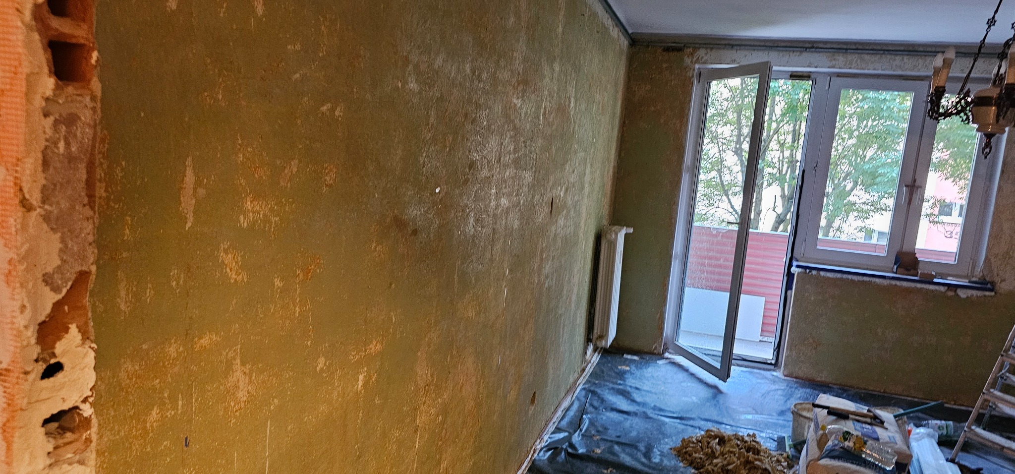 Pokój w trakcie remontu: ściany z resztkami starej farby, odkryta cegła, okno otwarte na oścież, zabezpieczona podłoga i narzędzia pracy.