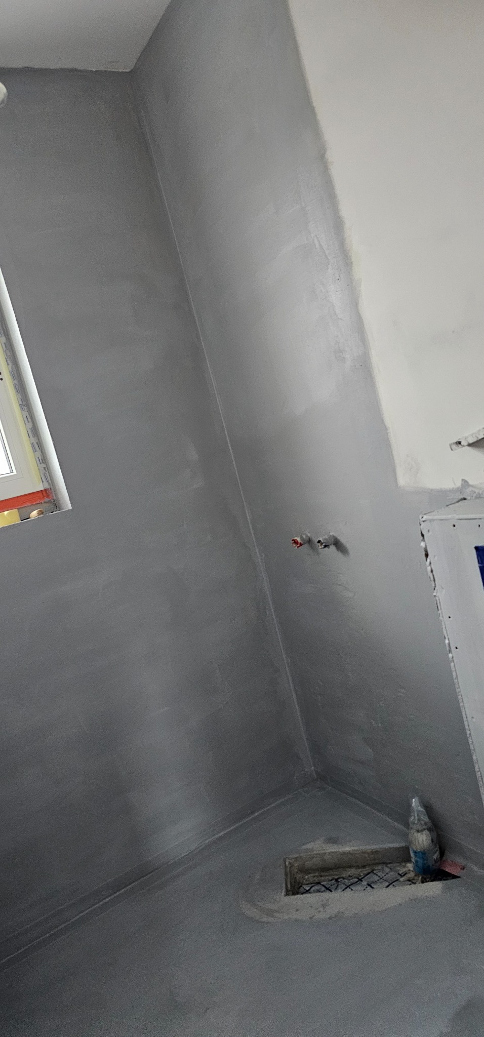 Świeżo wykończona, szara ściana i podłoga w łazience, widoczne fragmenty okna i instalacji wodnej, na podłodze resztki materiałów budowlanych obok kratki ściekowej.