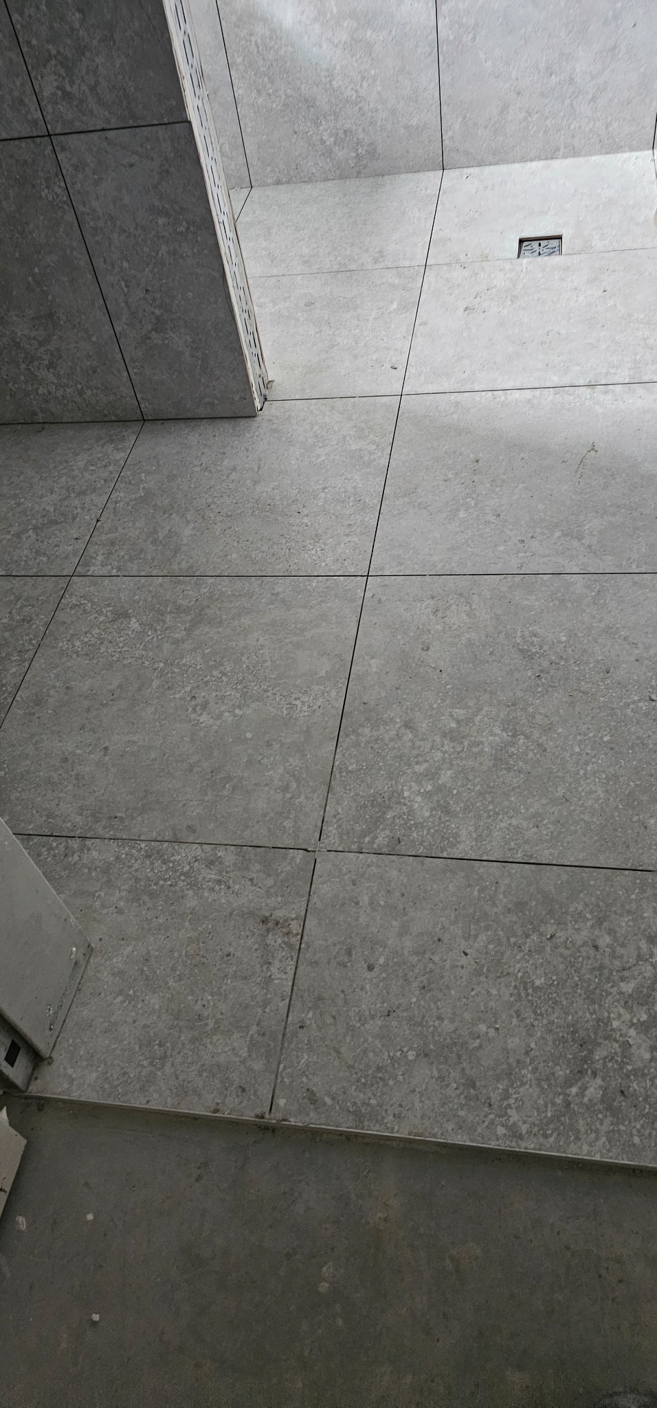 Ułożone szare, kwadratowe płytki podłogowe z widocznym odpływem liniowym i fragmentem progu, widziane pod kątem.