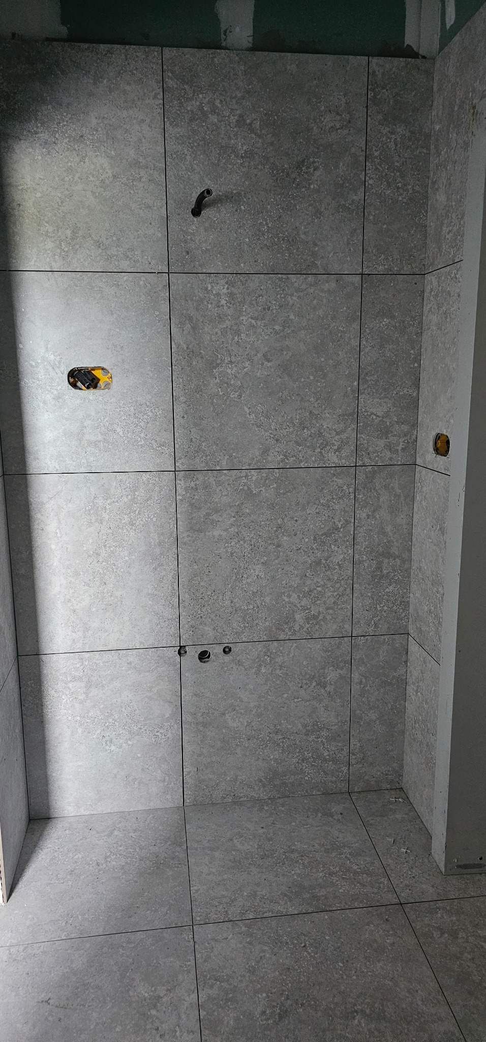 Ściana i podłoga w trakcie układania szarych, matowych płytek w łazience, widoczne otwory na instalacje i niedokończone wykończenie narożnika.