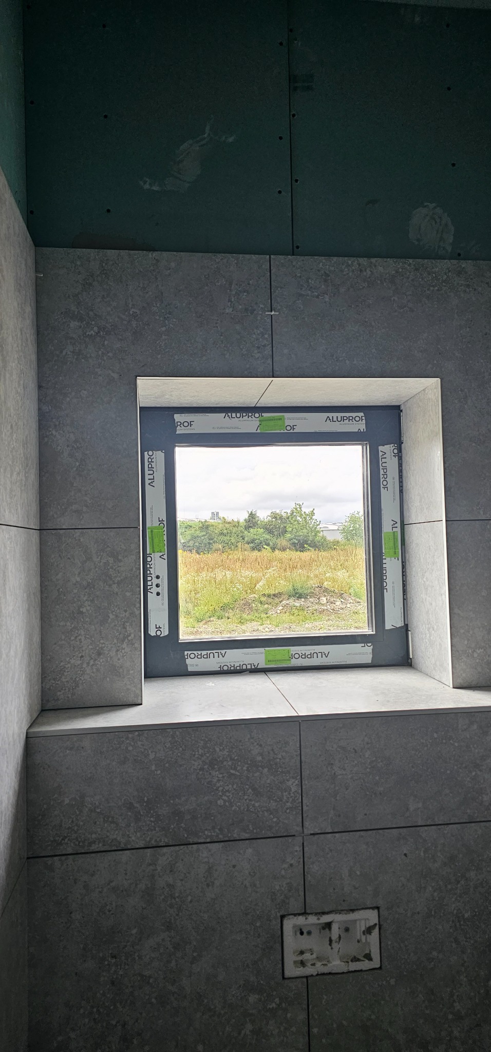 Szare płytki ceramiczne ułożone wokół okna z widokiem na zieleń, widoczne fugi i narożniki, na górze ściana z płyty kartonowo-gipsowej.