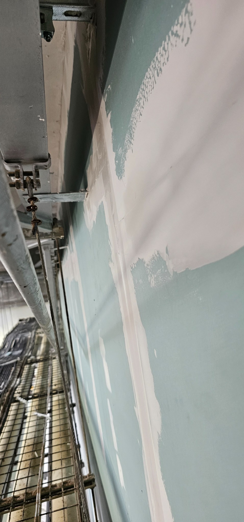 Nierównomiernie nałożona gładź szpachlowa na ścianie w trakcie prac wykończeniowych, widoczne metalowe elementy konstrukcyjne sufitu i oświetlenia.