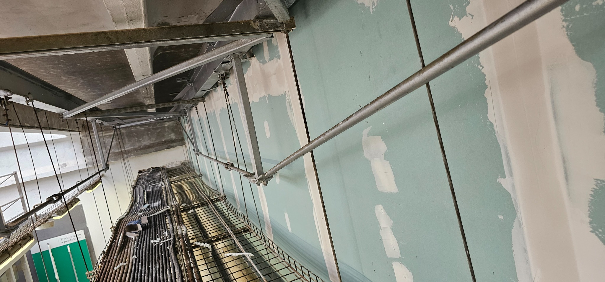 Ściana z płyt kartonowo-gipsowych w trakcie szpachlowania, widoczne metalowe konstrukcje pod sufitem i koryta kablowe.